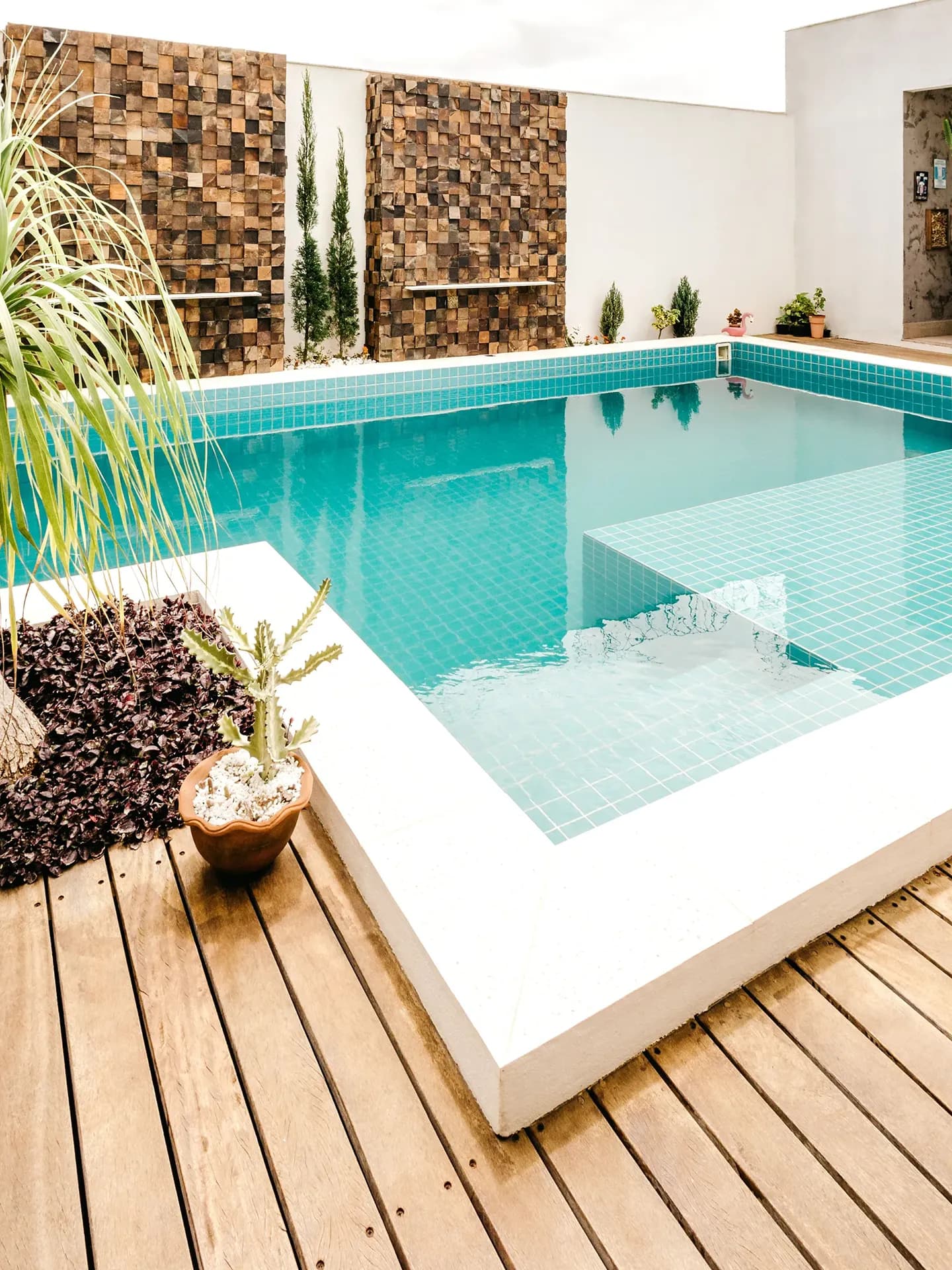 A modern courtyard swimming pool with turquoise mosaic tiles and crisp white coping, flanked by a natural wood deck, a potted cactus, and prominent textured wooden block feature walls