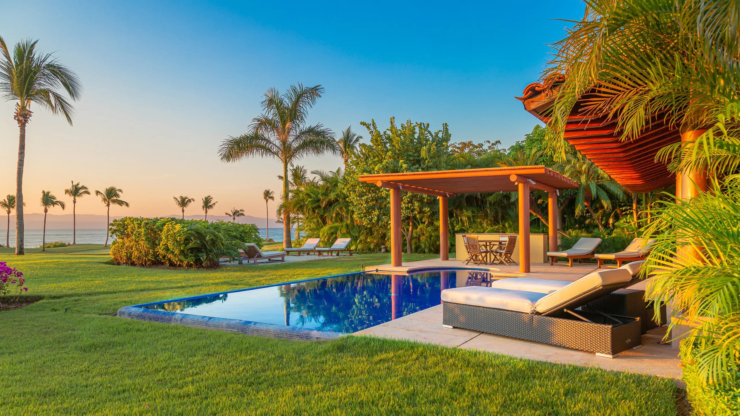 An elegant luxury villa terrace at sunset, showcasing a clear blue swimming pool, wooden pergolas, comfortable lounge chairs, and palm trees on a manicured lawn overlooking the ocean.