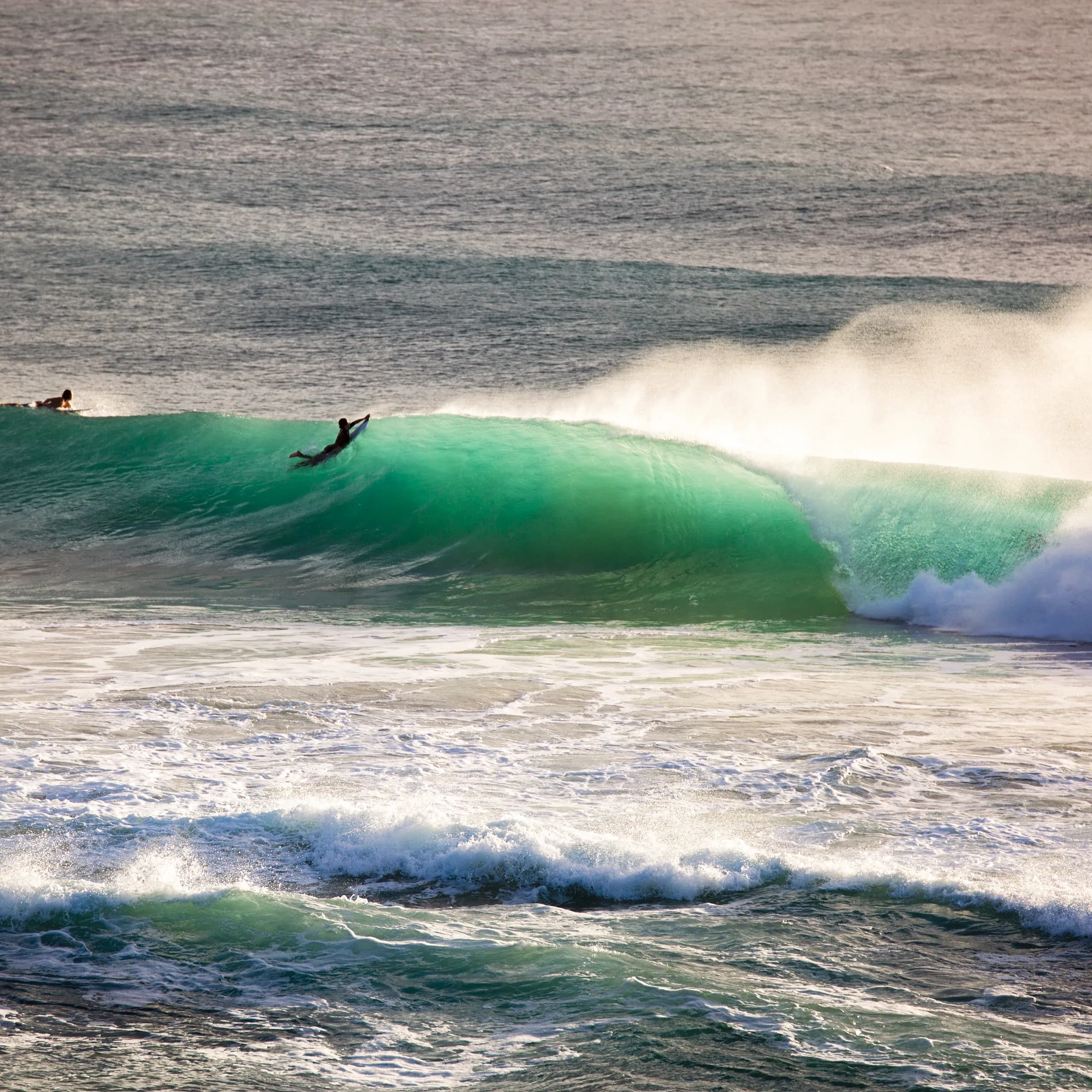 A surfer paddling down a translucent, bright turquoise cresting ocean wave, with sea spray catching the golden afternoon light.