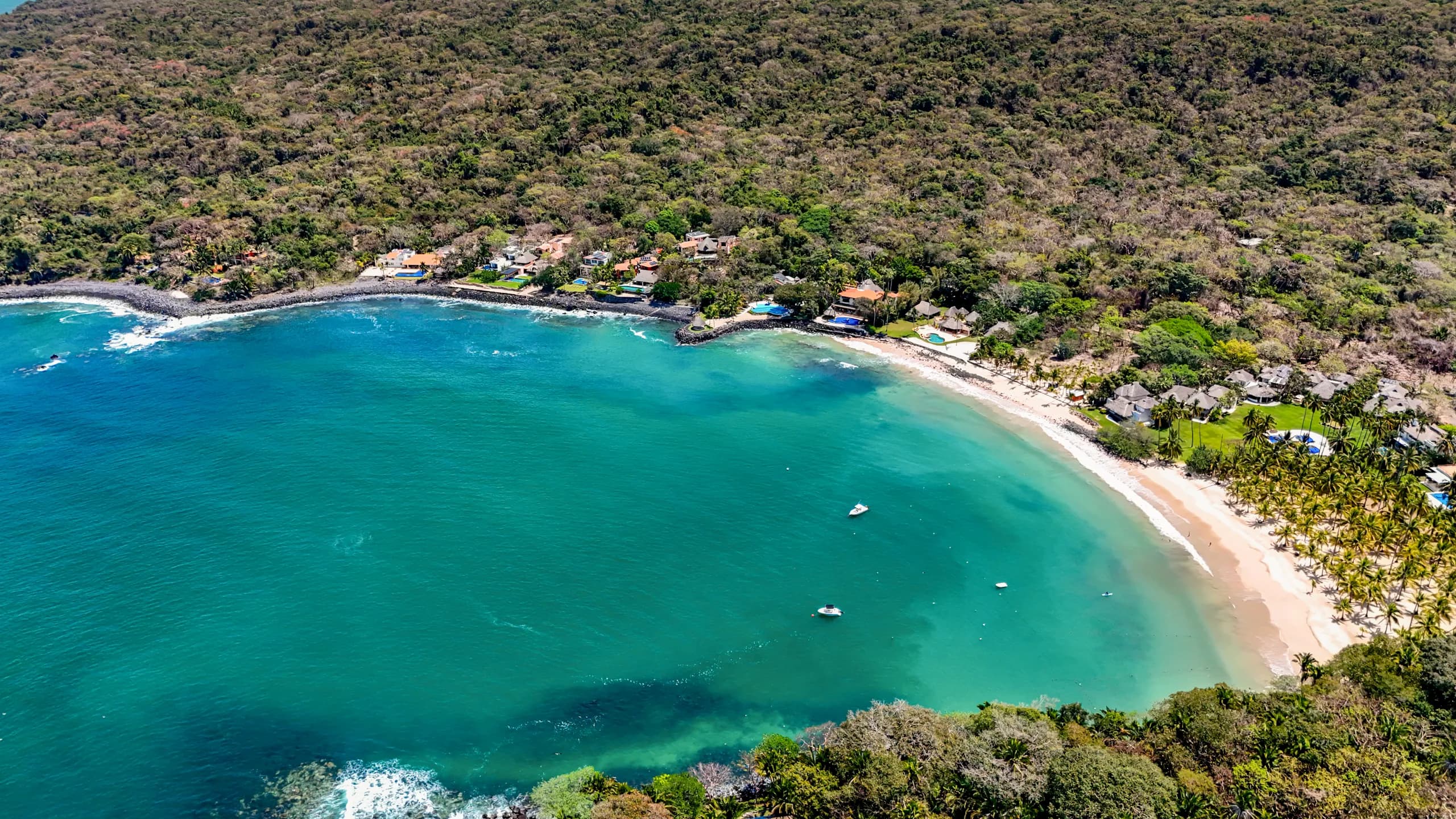 An aerial drone view of Chacala bay, showing a crescent-shaped sandy beach with turquoise water and a few small boats anchored, bordered by a dense green jungle forest and coastal residences.