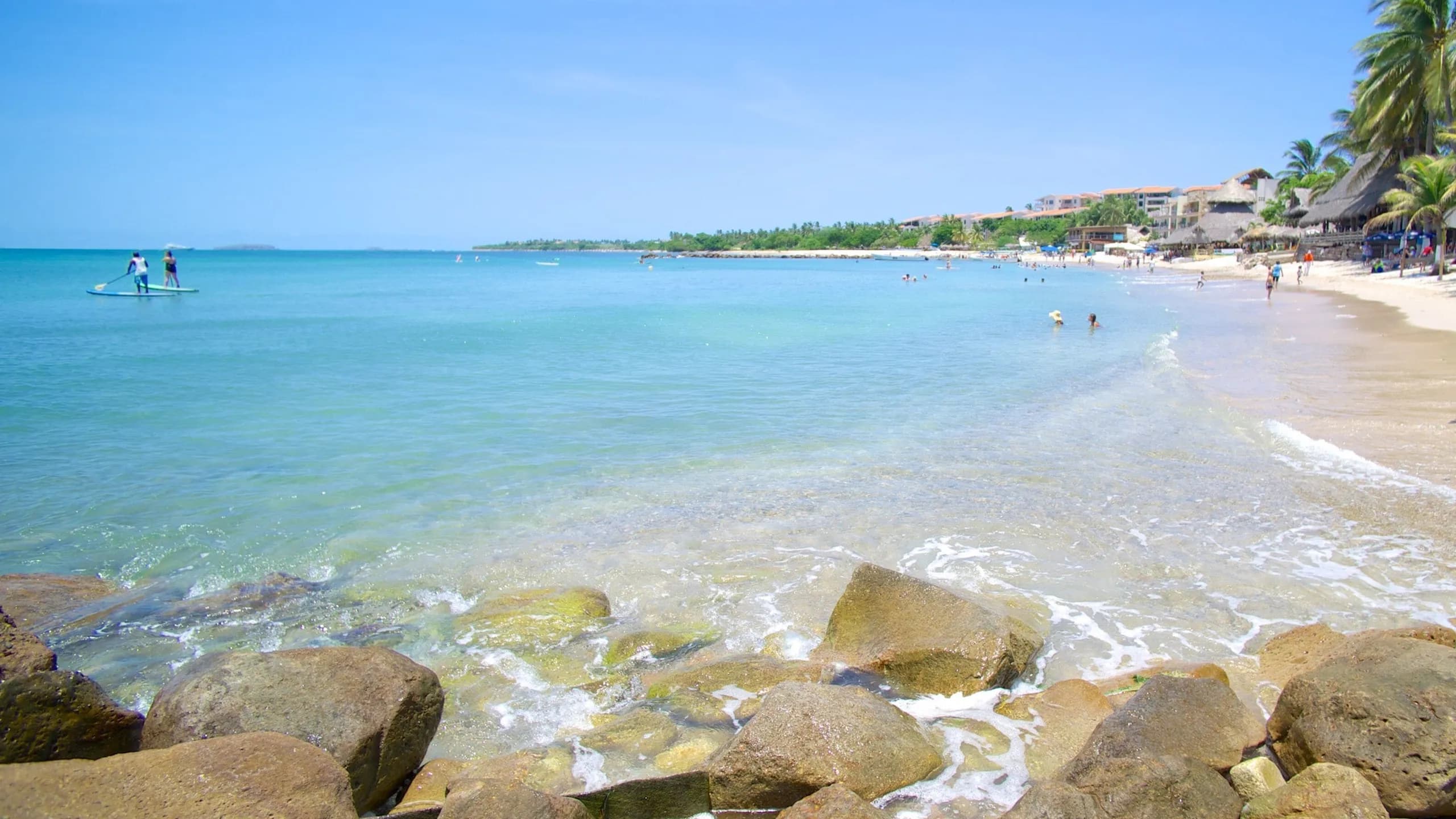 A wide view of El Anclote beach featuring crystalline, calm turquoise water with rocks in the immediate foreground, paddleboarders out on the sea, and people walking on the sandy shore lined with palm trees and palapa buildings under a clear sky.