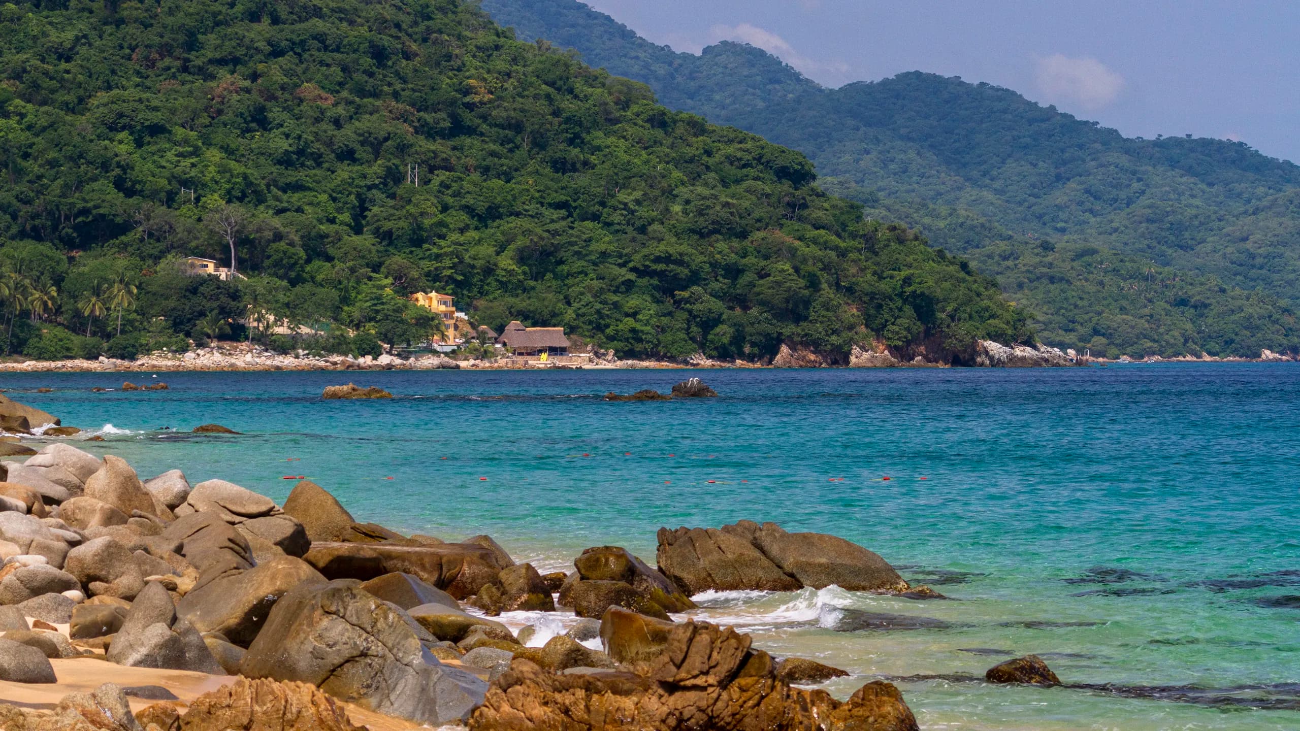 A scenic landscape view of Playa Caballos along the hiking trail to Las Animas, featuring a rocky shoreline with large smooth boulders in the foreground, vibrant turquoise water, and lush green tropical mountains with scattered coastal homes in the distance.