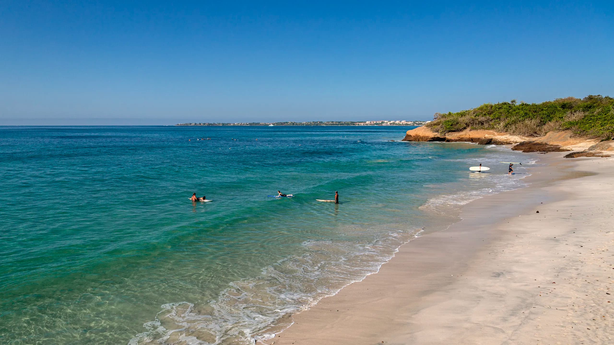 A wide sunny view of La Lancha beach, showing surfers scattered in the clear turquoise ocean water and others walking along the sandy shore, bordered by a rocky, tree-covered point under a vast blue sky.