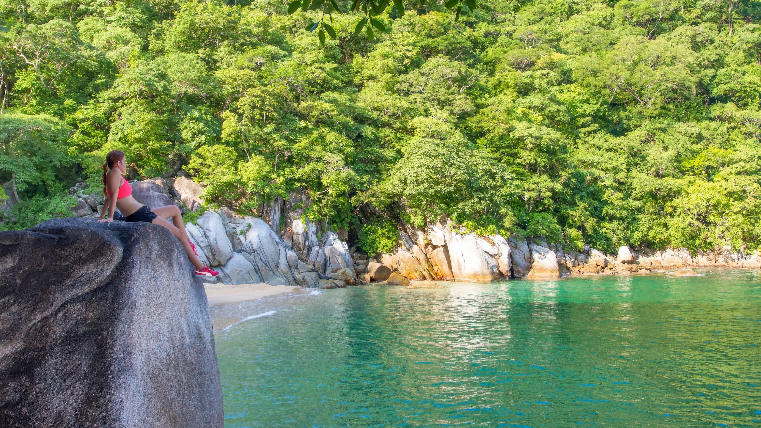 A woman in athletic wear sitting atop a massive dark boulder overlooking the clear emerald green water of Colomitos cove, with a small sandy shore and dense tropical jungle-covered cliffs in the background.