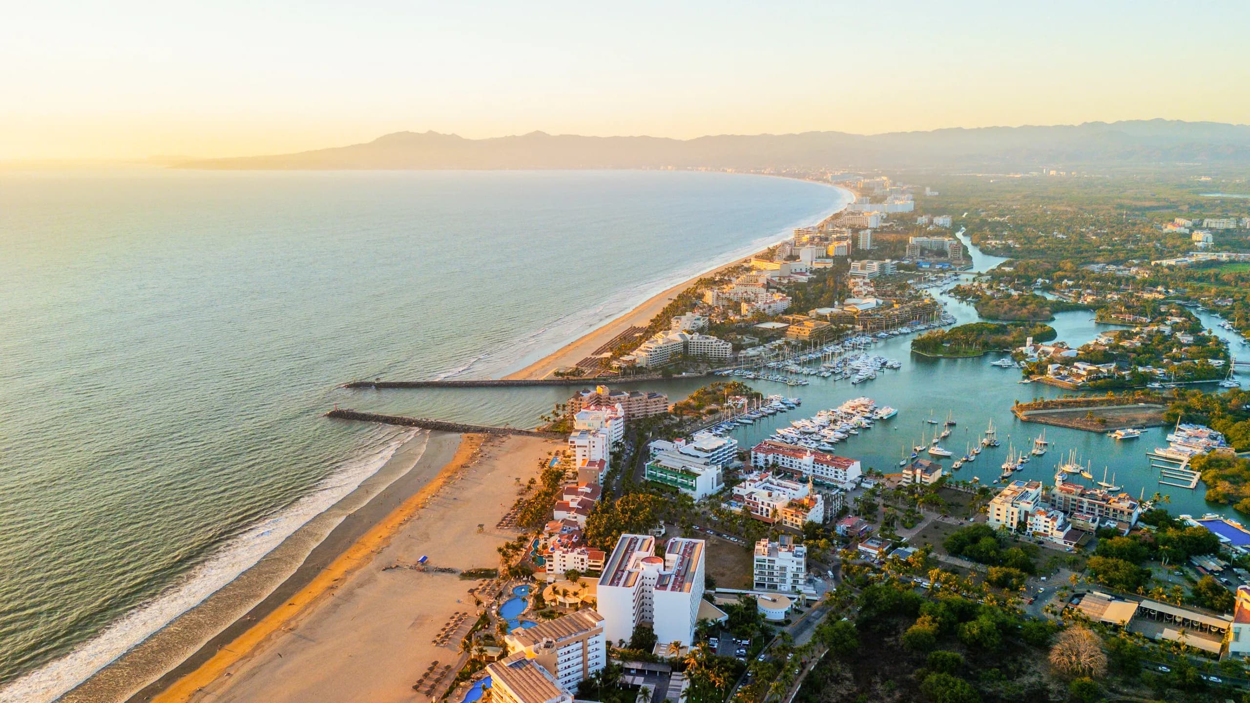 A high aerial drone sunset view over the Nuevo Nayarit coastline and marina, showcasing long sandy beaches separated by stone jetties, a harbor filled with numerous white boats and yachts, and coastal resorts stretching toward distant mountains.