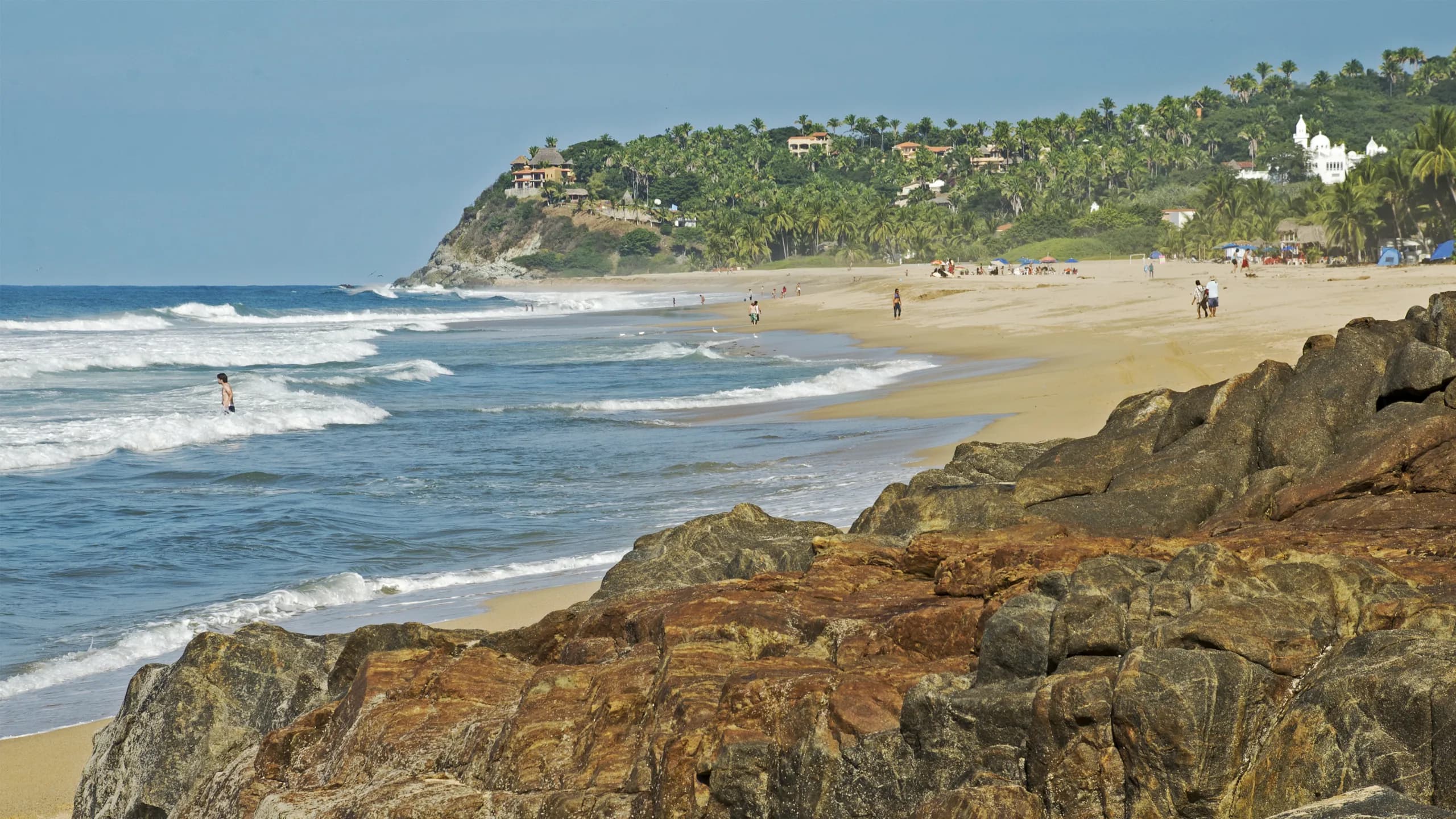 A scenic view of San Pancho beach looking down the coastline, featuring large brown rocks in the foreground, rolling ocean waves with a person wading in the surf, and a long sandy beach bordered by tropical green hills and palm trees under a blue sky.