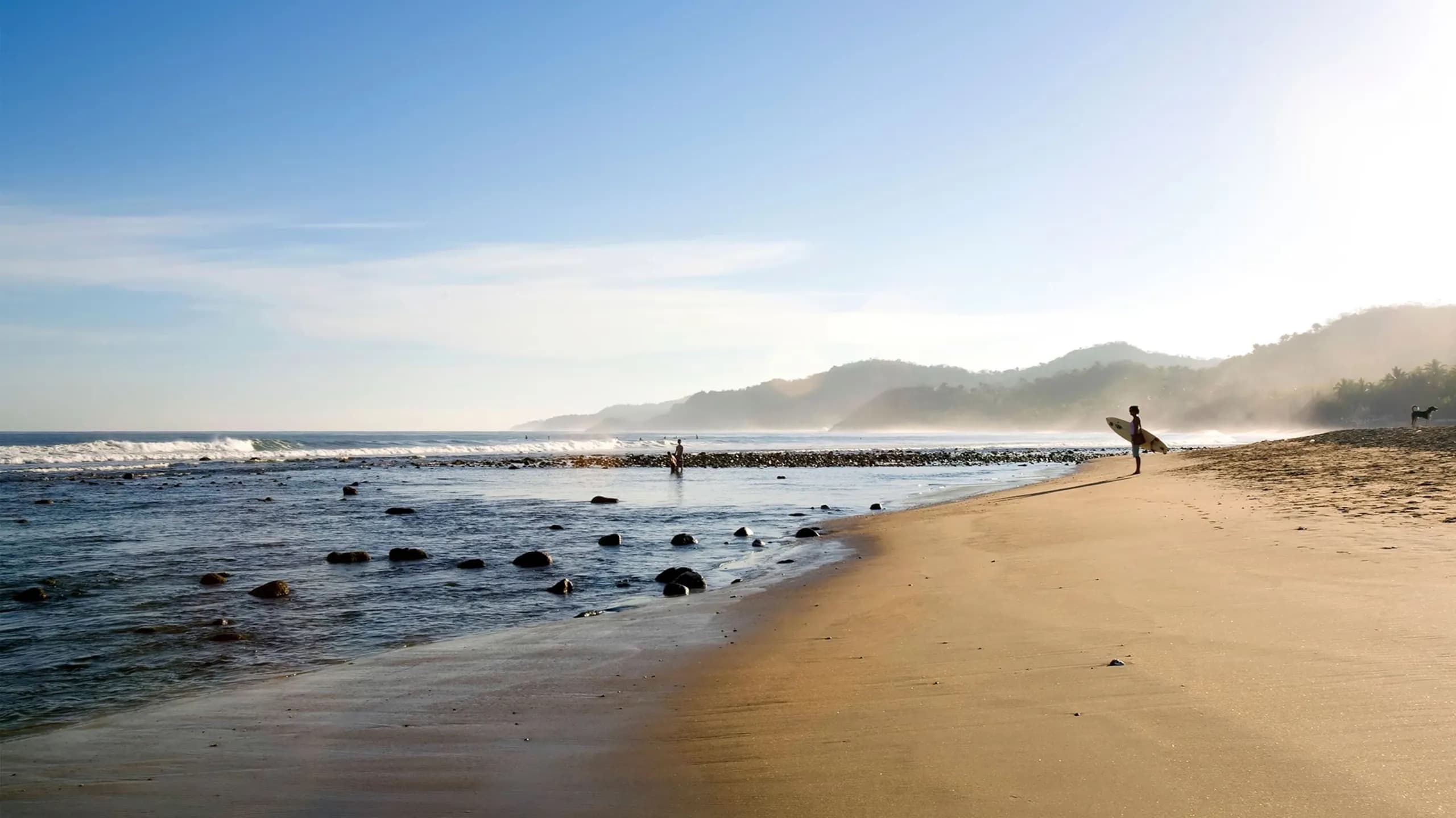 A wide, low-angle shot of a serene beach in Sayulita during early morning, featuring a surfer standing with a board on the sandy shore looking out at gentle ocean waves breaking over rocks, with hazy mountains in the distant background.