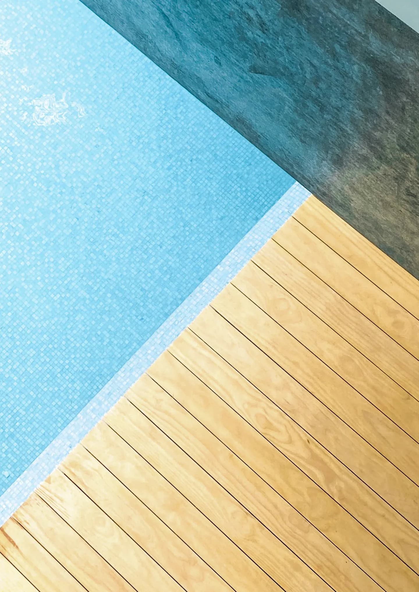 A minimalist, high-angle geometric composition showing a natural wood deck meeting the light blue mosaic tile of a swimming pool and a dark gray concrete pool deck.