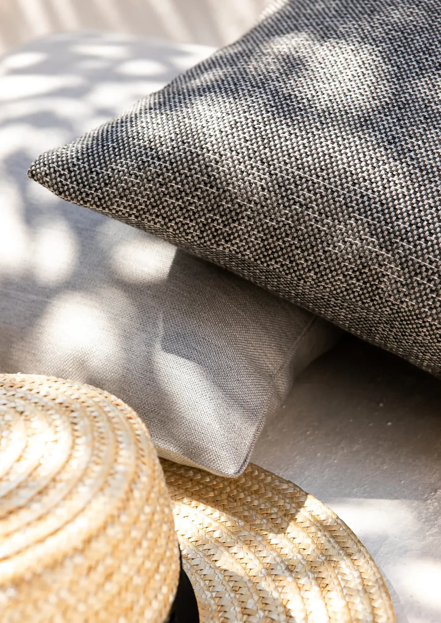 A close-up shot of two outdoor pillows showing rich fabric textures in gray and woven black-and-white patterns, placed next to a woven straw hat under soft shadows.