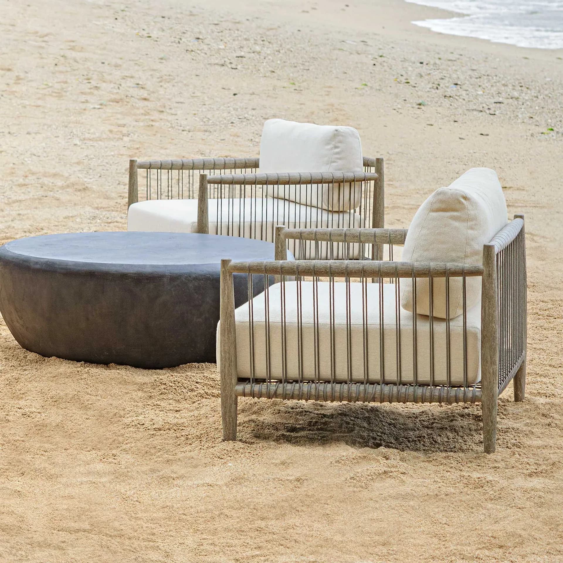 Two luxury lounge chairs with beige upholstered cushions and rope-wrapped frames set directly on beach sand next to a low, dark stone coffee table near the ocean surf.