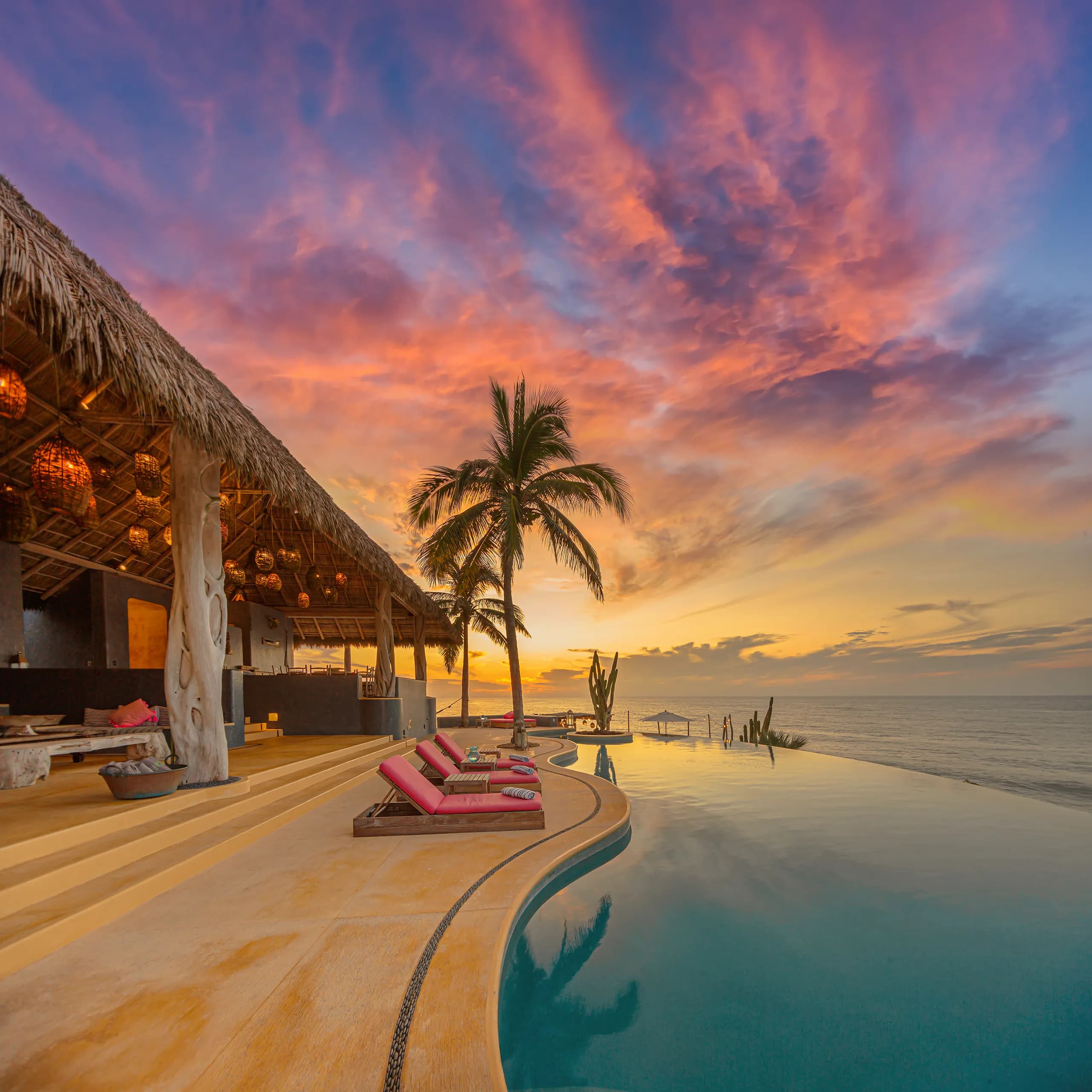 Luumhaus beachfront residence showcasing a luxury infinity pool, palapa lounge architecture, and sunbeds under a vibrant orange and pink sunset sky