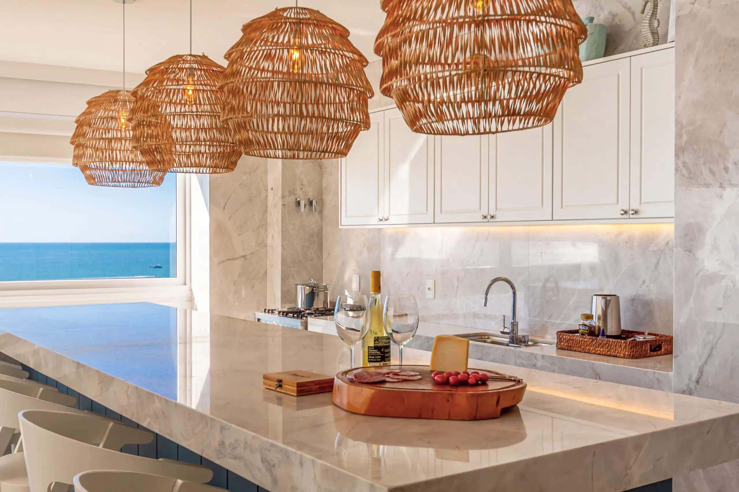 A luxury coastal kitchen featuring a polished genuine quartzite island and matching backsplash, accented by large woven rattan pendant lights and an open view of the blue ocean.