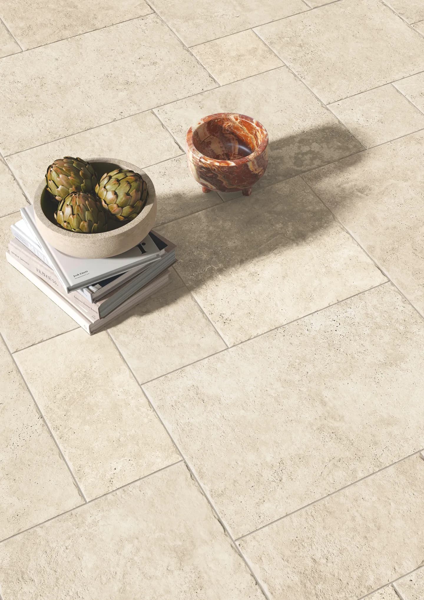 An overhead view of a textured travertine tile floor in a warm, cream tone, styled with a stone bowl of artichokes stacked on design books and a small marbled stone bowl.