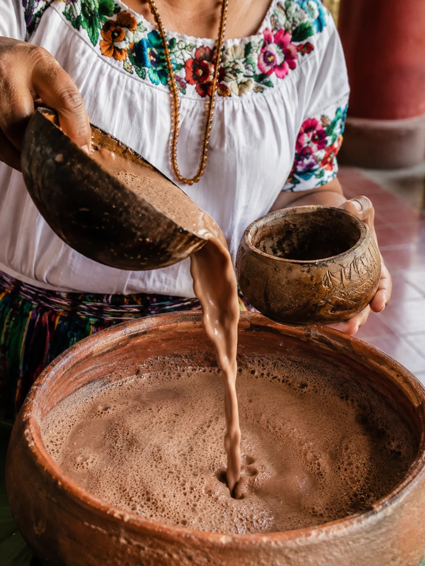 An Indigenous woman wearing a traditional floral embroidered white blouse pouring frothy <span class="italic">tanchuca</span> drink from a <span class="italic">jícara</span> gourd bowl into a large clay basin.