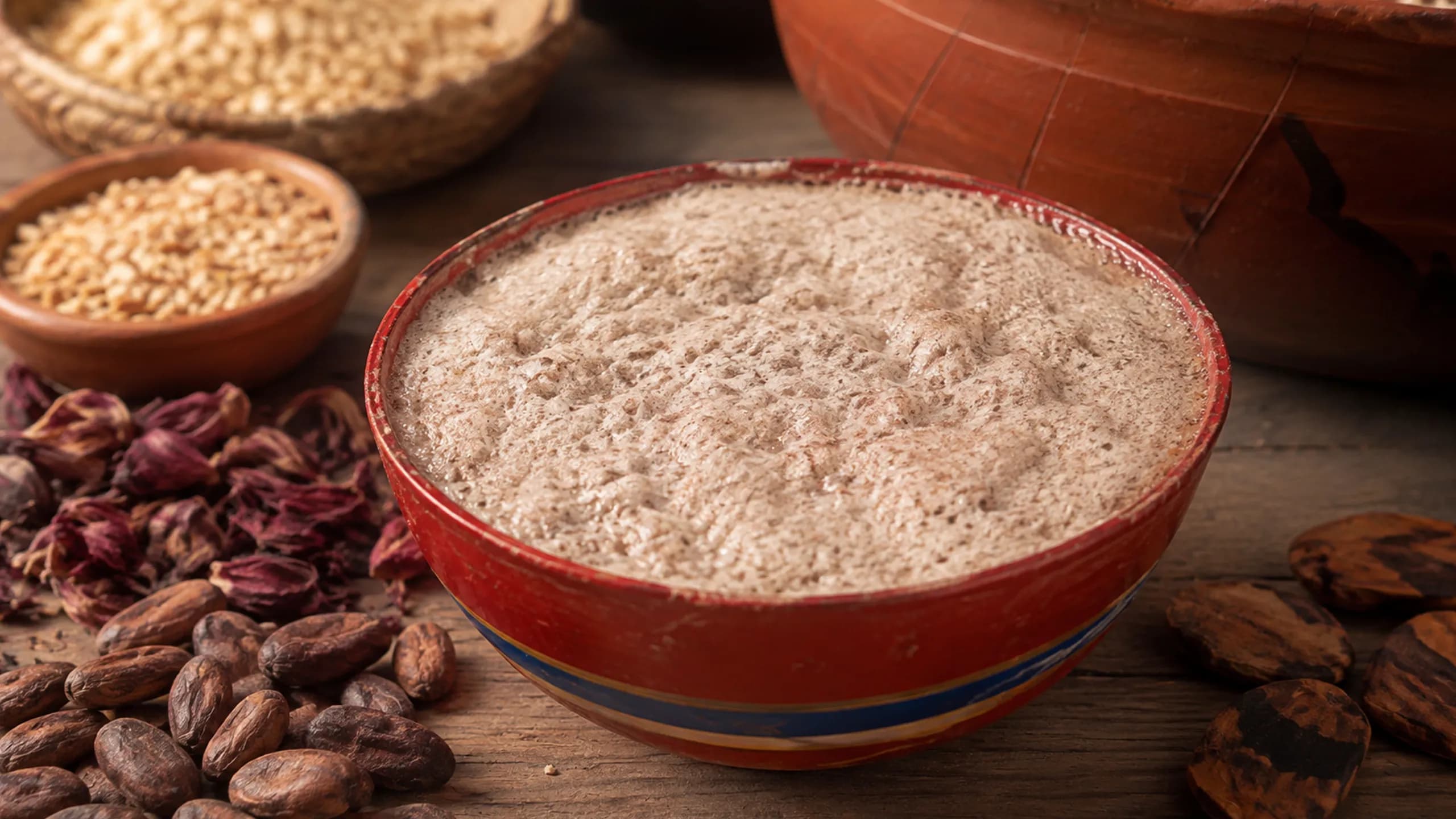 A hand-painted traditional bowl overflowing with thick, pale frothy <span class="italic">tejate</span> drink, surrounded by cacao beans, <span class="italic">mamey</span> seeds (pixtle), rosita de cacao flowers, and corn in small clay vessels.