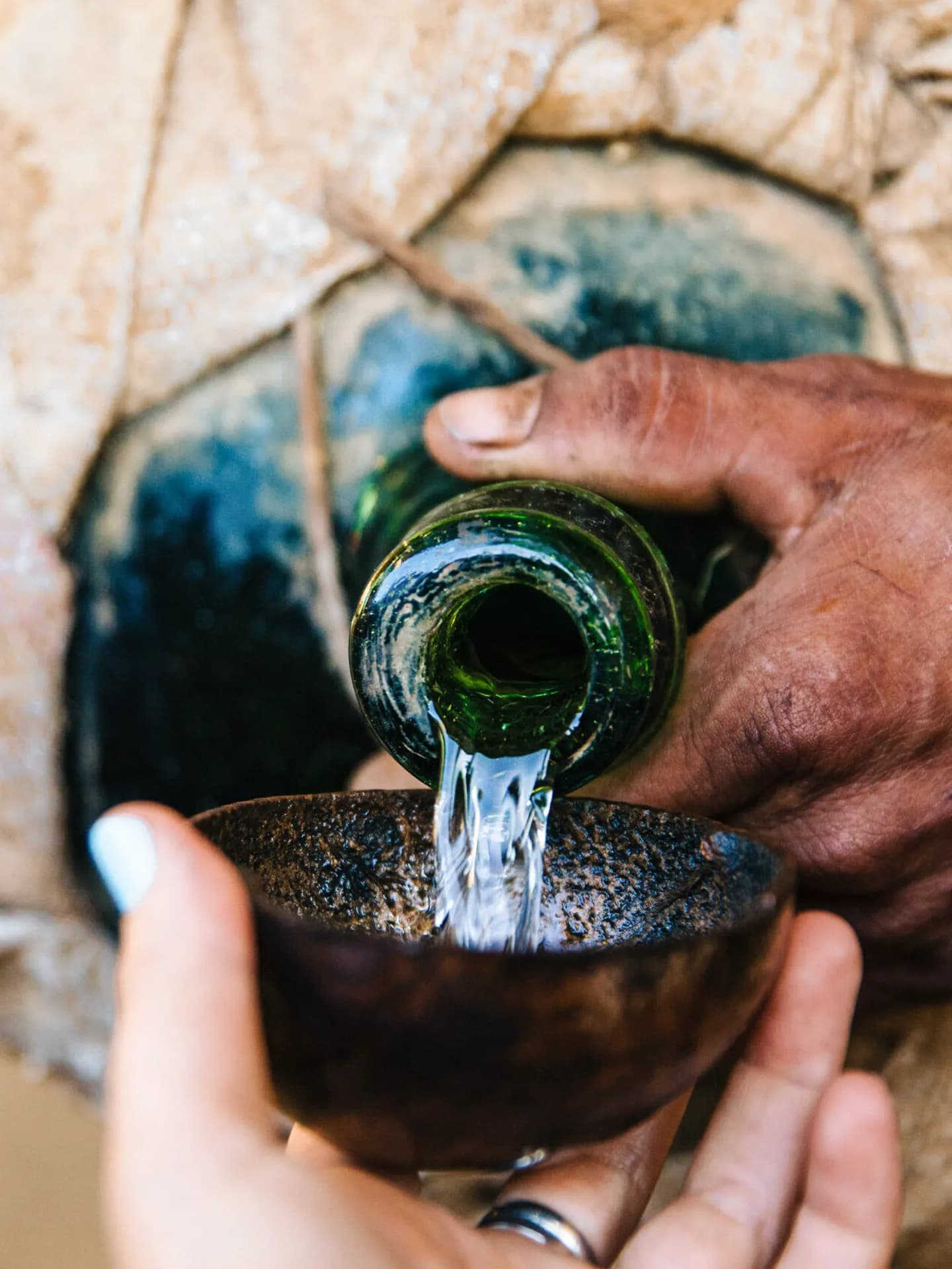 Una toma de cerca de raicilla transparente siendo servida desde una botella de vidrio verde hacia una jícara tradicional de cuastecomate de textura oscura sostenida con la mano.