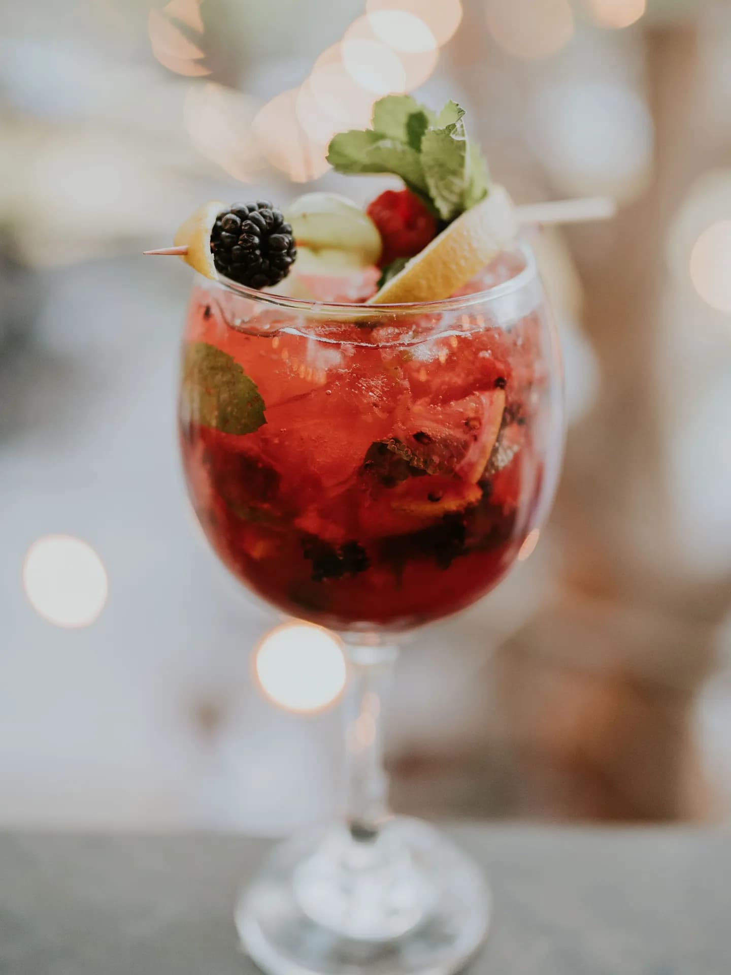 A vibrant red raicilla cocktail served in a stemmed balloon glass with crushed ice, garnished with fresh blackberries, citrus slices, berries on a skewer, and a mint sprig, against a warm bokeh background.