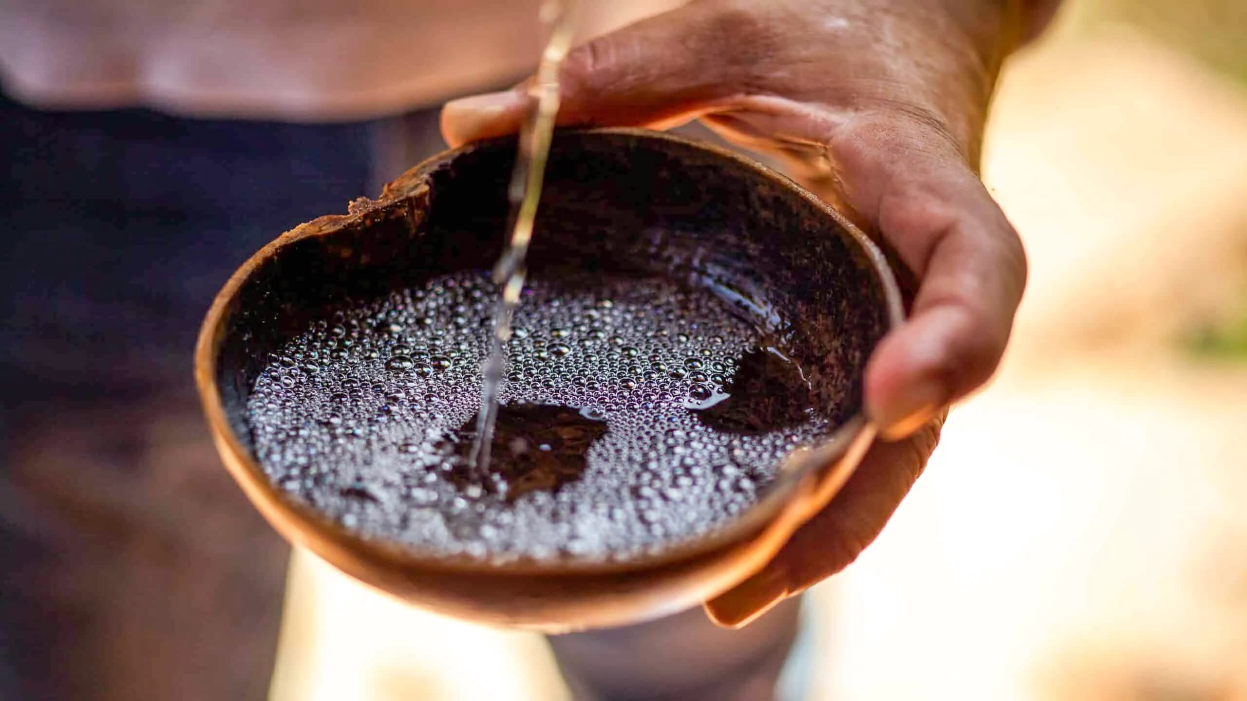 A close-up view of freshly distilled, clear Raicilla being poured into a cuastecomate jícara, creating delicate pearls or bubbles on the surface.
