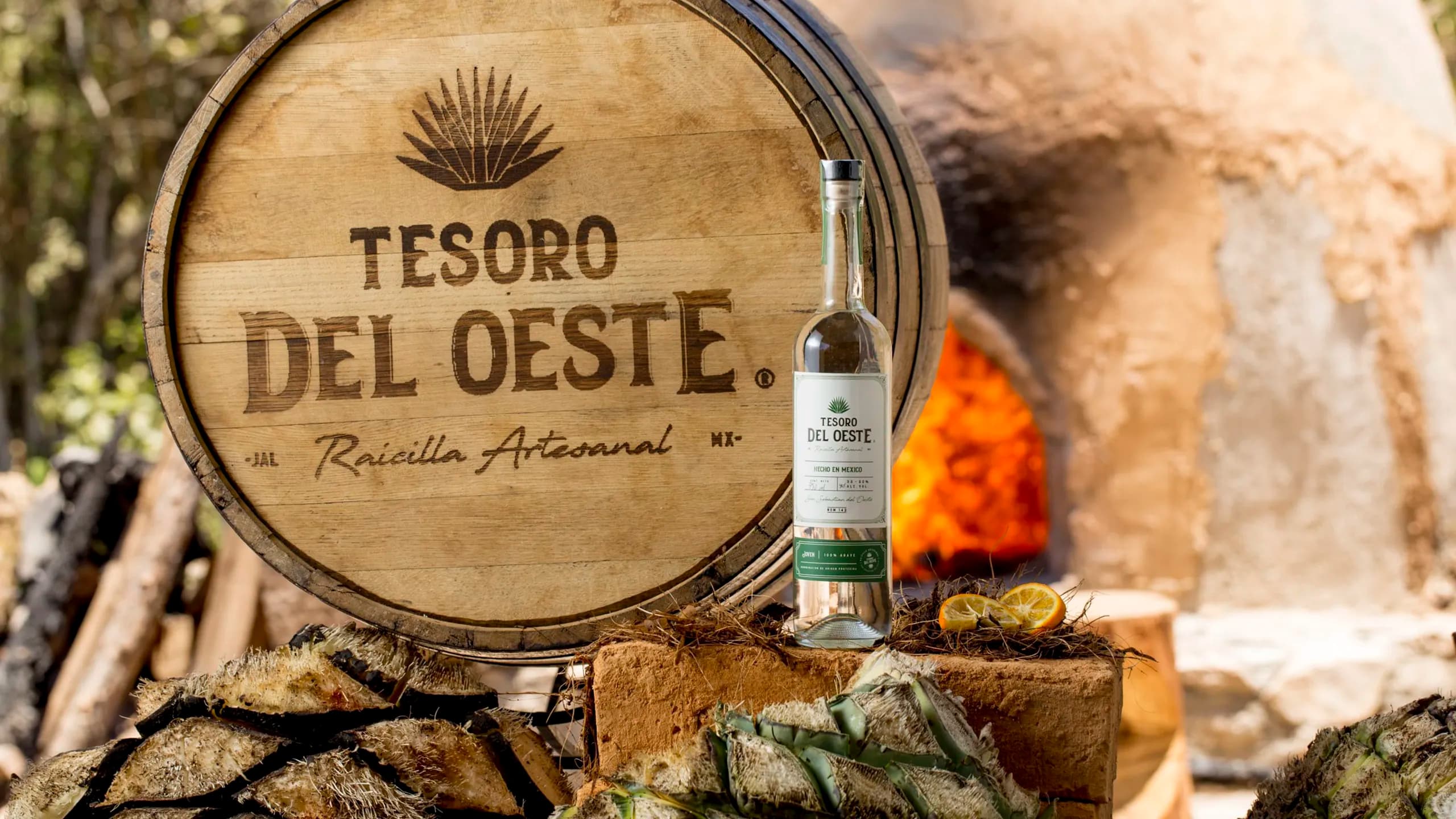 Una botella transparente de Raicilla Artesanal Tesoro del Oeste junto a una tapa de barril de madera con el logotipo de la marca, rodeada de piñas de agave cosechadas y un horno de adobe encendido al fondo.
