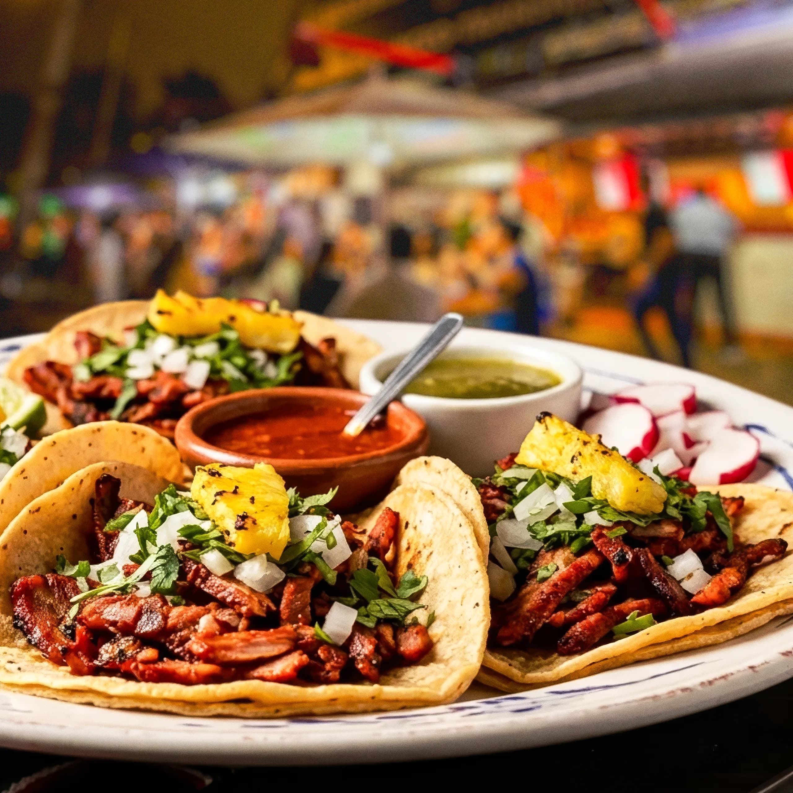 A plate of freshly prepared tacos al pastor topped with charred marinated pork, pineapple slices, chopped onions, and cilantro, served alongside red and green salsas and sliced radishes.