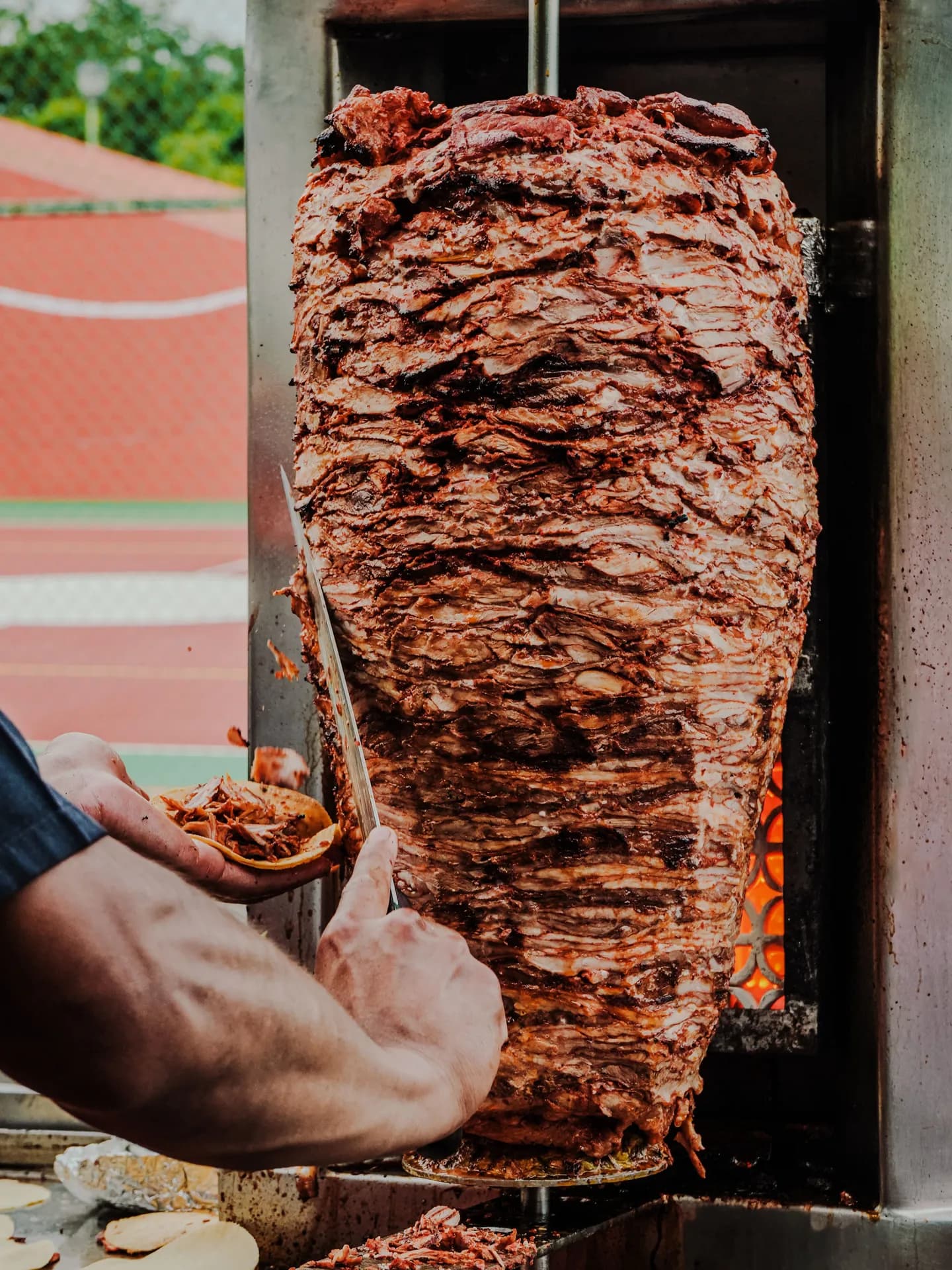 A taquero using a long knife to slice thin ribbons of charred, marinated pork off a large vertical trompo directly into a warm tortilla.