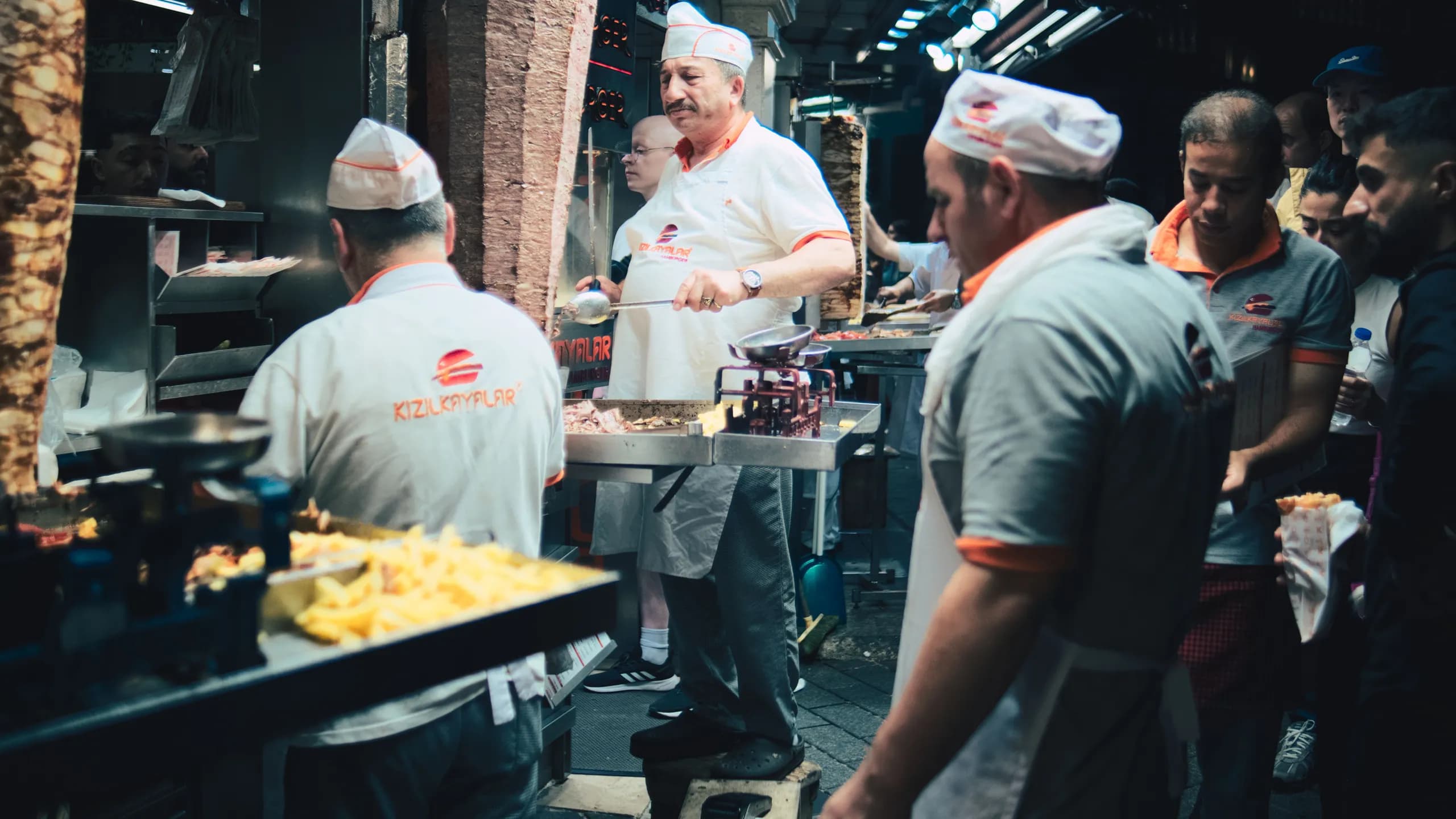 Middle Eastern chefs in white uniforms preparing shawarma meat on large vertical spits at a busy street food counter.