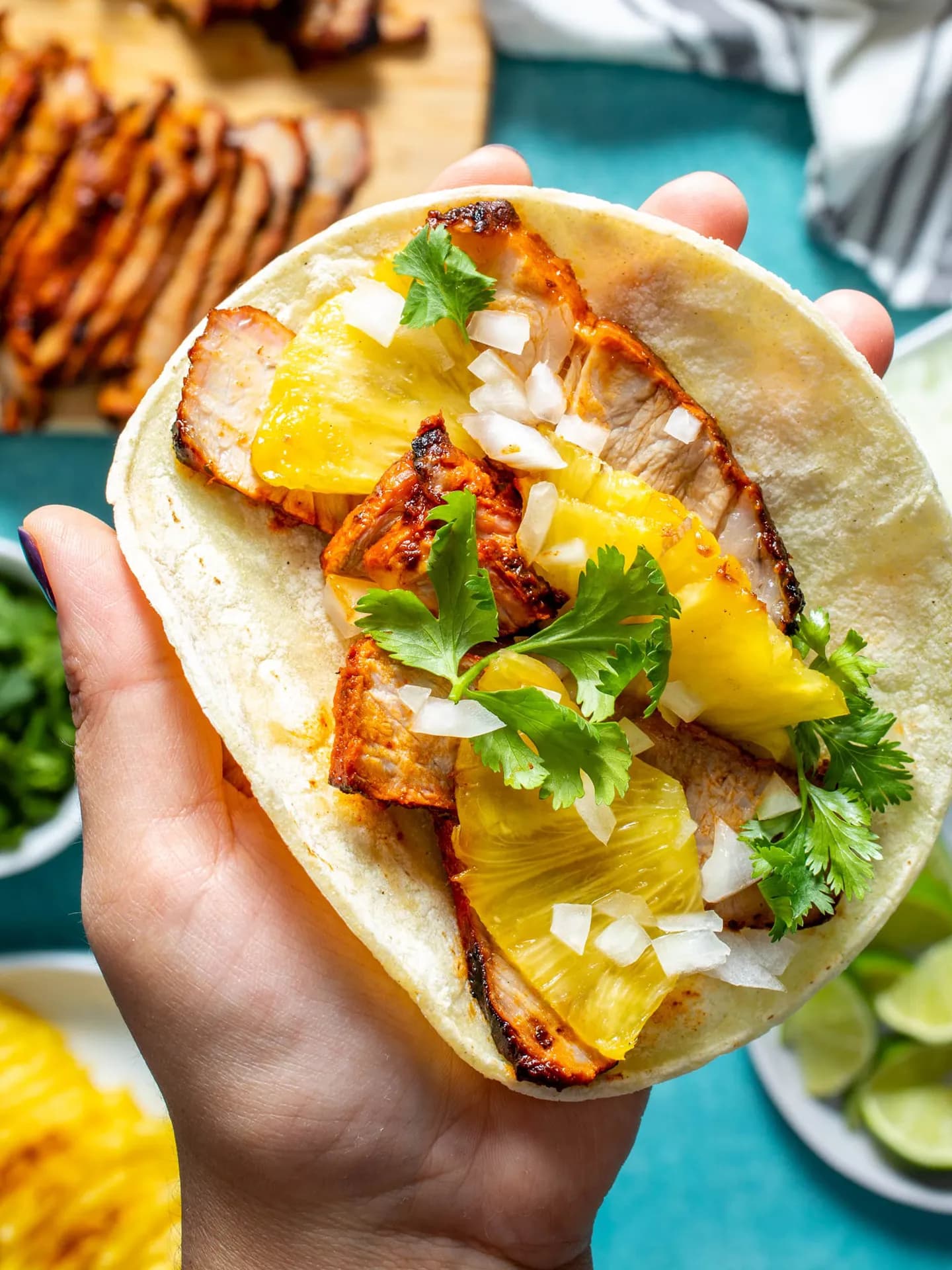 A close-up overhead shot of a hand holding an open corn tortilla taco loaded with thin slices of marinated pork al pastor, roasted pineapple slices, diced white onion, and fresh cilantro leaves.