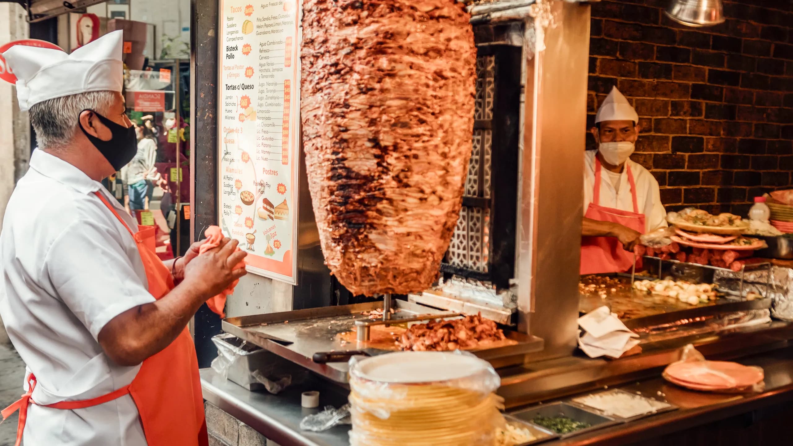 Two taqueros wearing traditional white chef hats and aprons standing by a large vertical meat spit at a taquería counter with a menu board in the background.