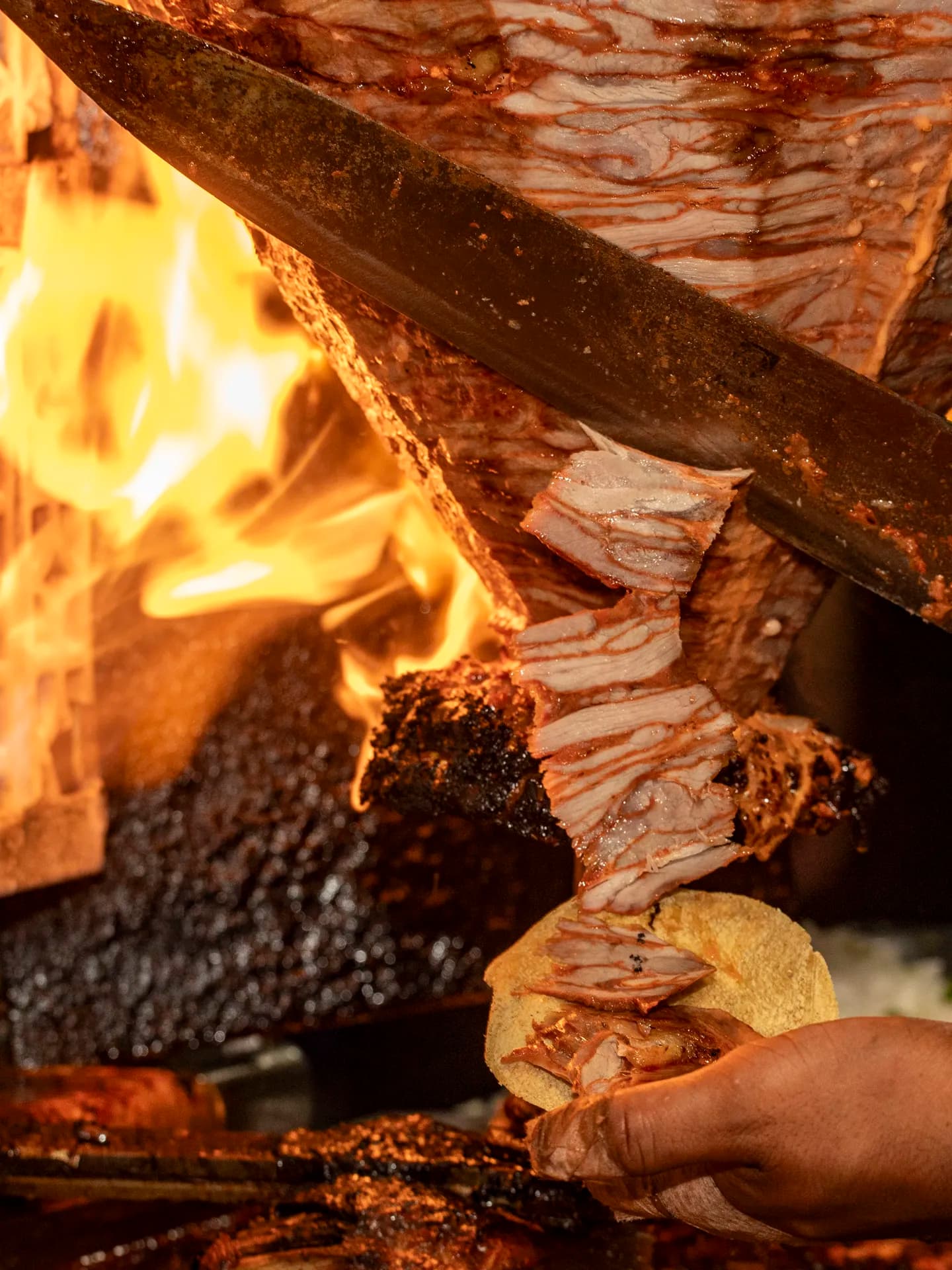 A close-up action shot of a long knife carving succulent ribbons of marinated pork off a trompo right in front of open flames onto a warm tortilla.