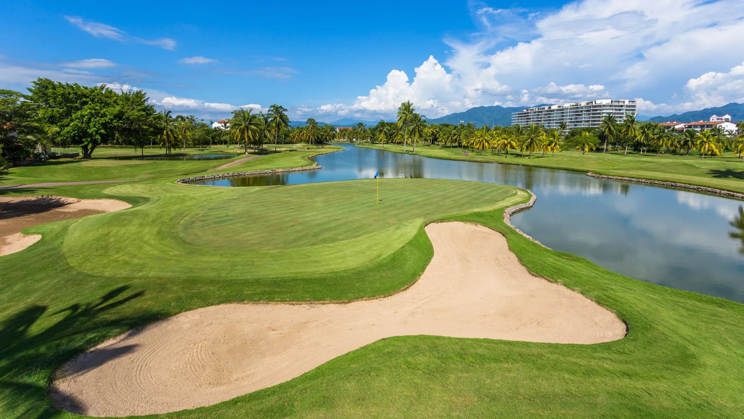 Un green impecable resguardado por un bunker de arena y un lago Serpenteante en Marina Vallarta Golf Club, con palmeras y arquitectura residencial de fondo.