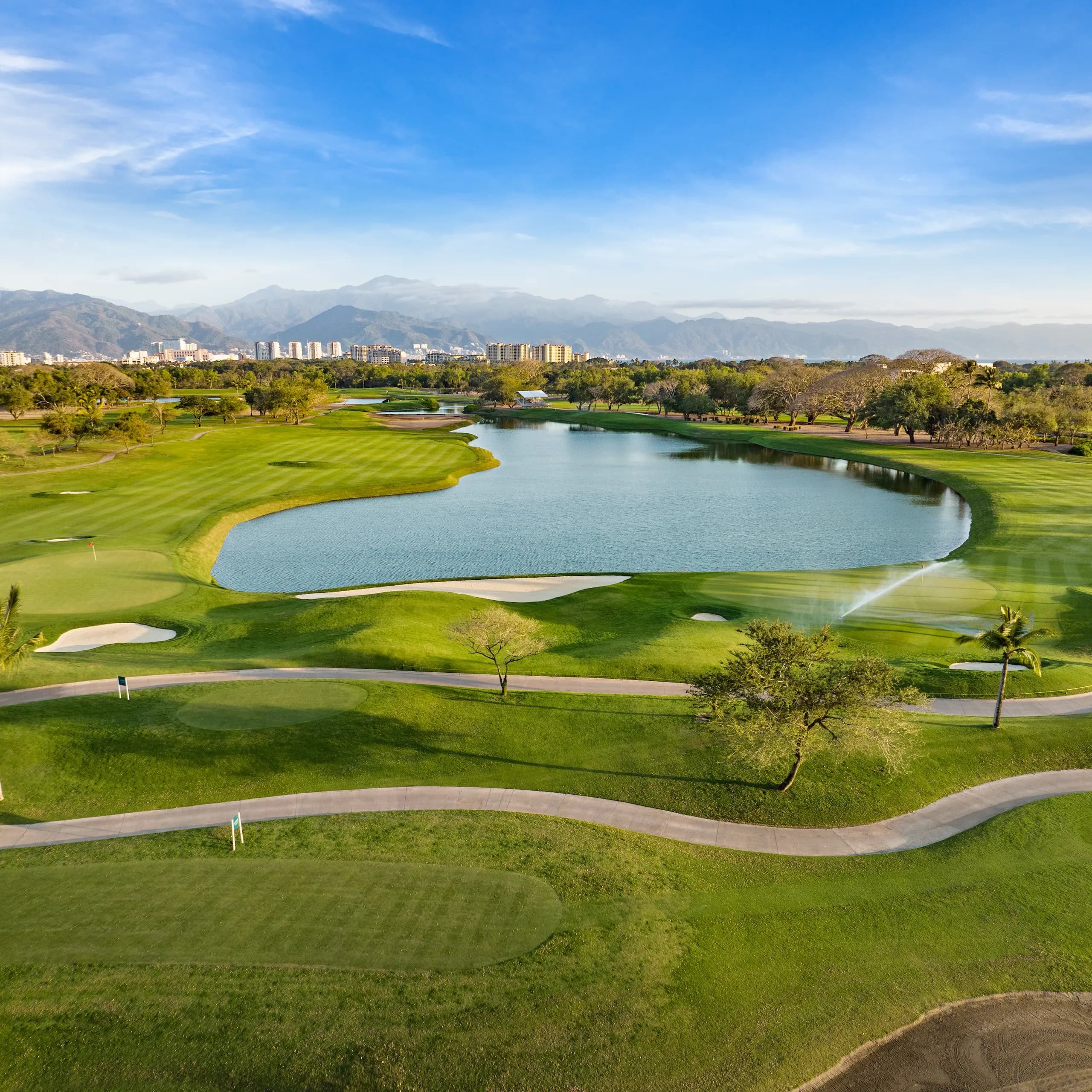 Vista aérea de un exuberante campo de golf verde en Vidanta, que destaca un gran lago, caminos sinuosos para carritos, árboles esparcidos y el horizonte distante con un pintoresco fondo montañoso.