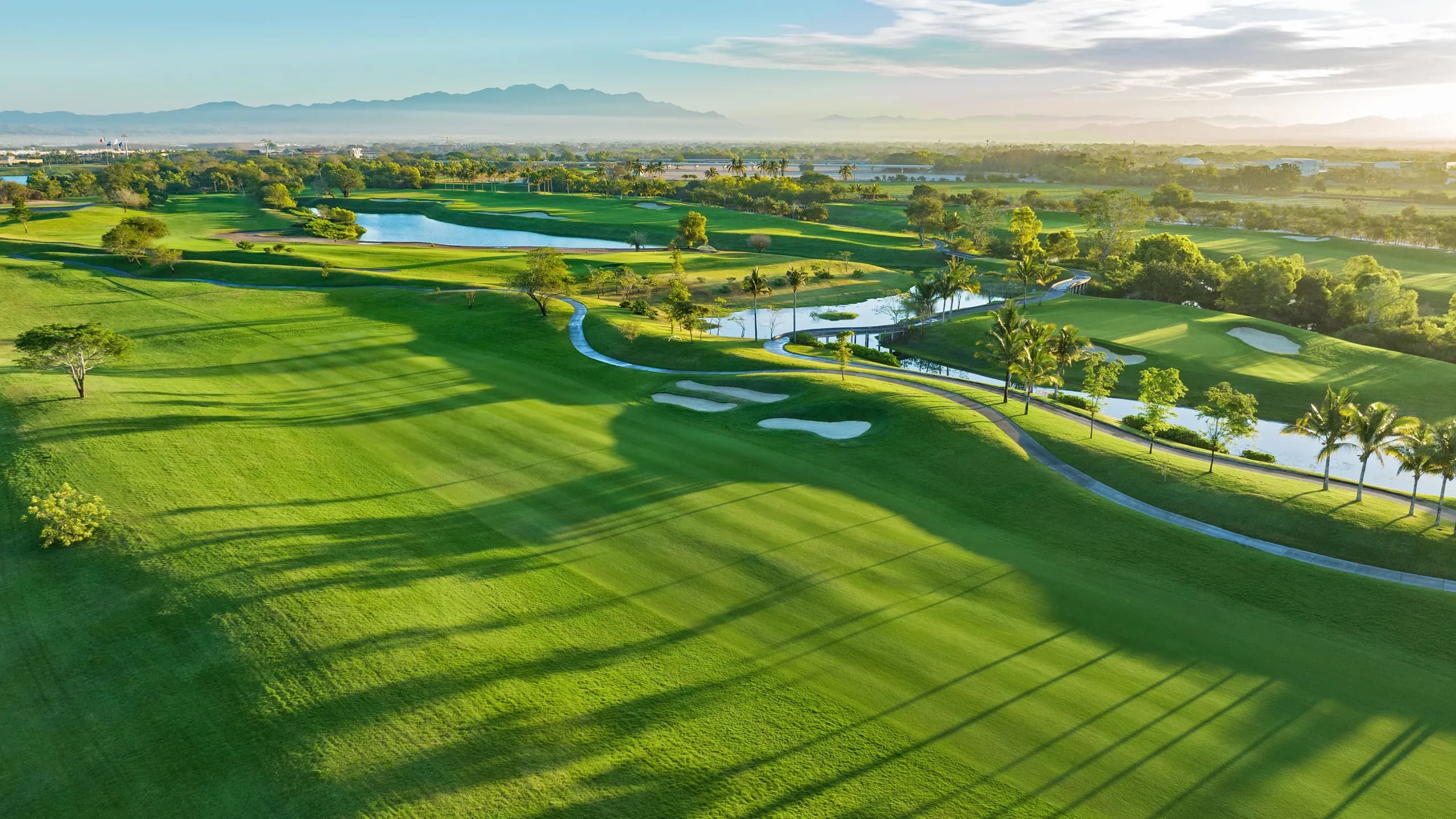 Vista aérea del campo de golf Vidanta Vallarta mostrando los fairways de paspalum, sus lagos y bunkers, con las montañas de la Sierra Madre al fondo.