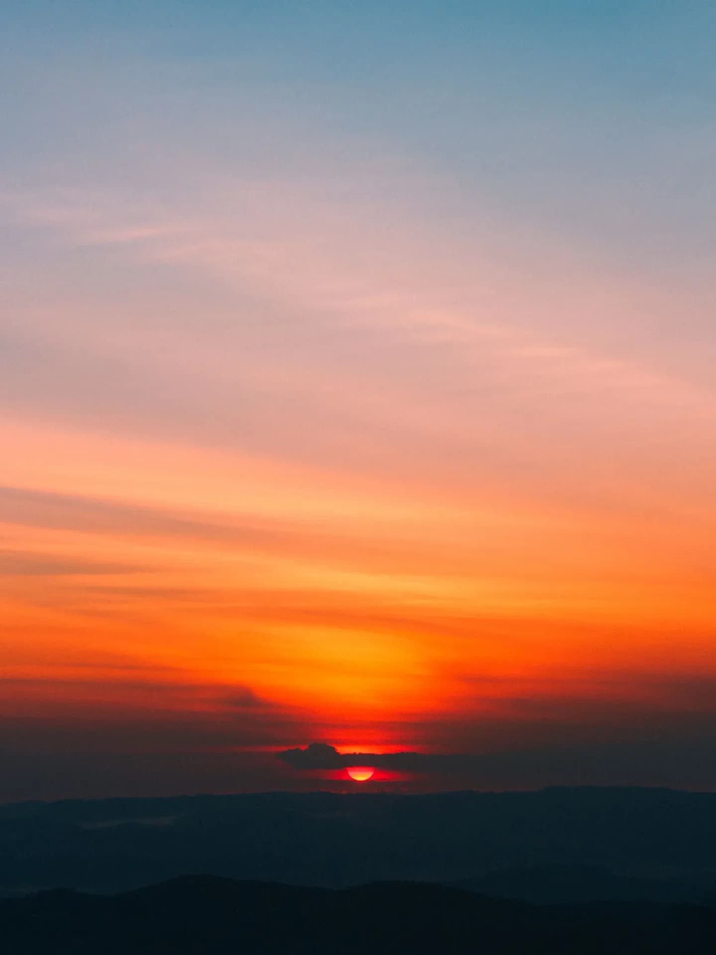 A breathtaking vertical sunset over distant dark mountain silhouettes, with a fiery red sun dipping below thin clouds and a gradient sky shifting from brilliant orange to soft pastel blue