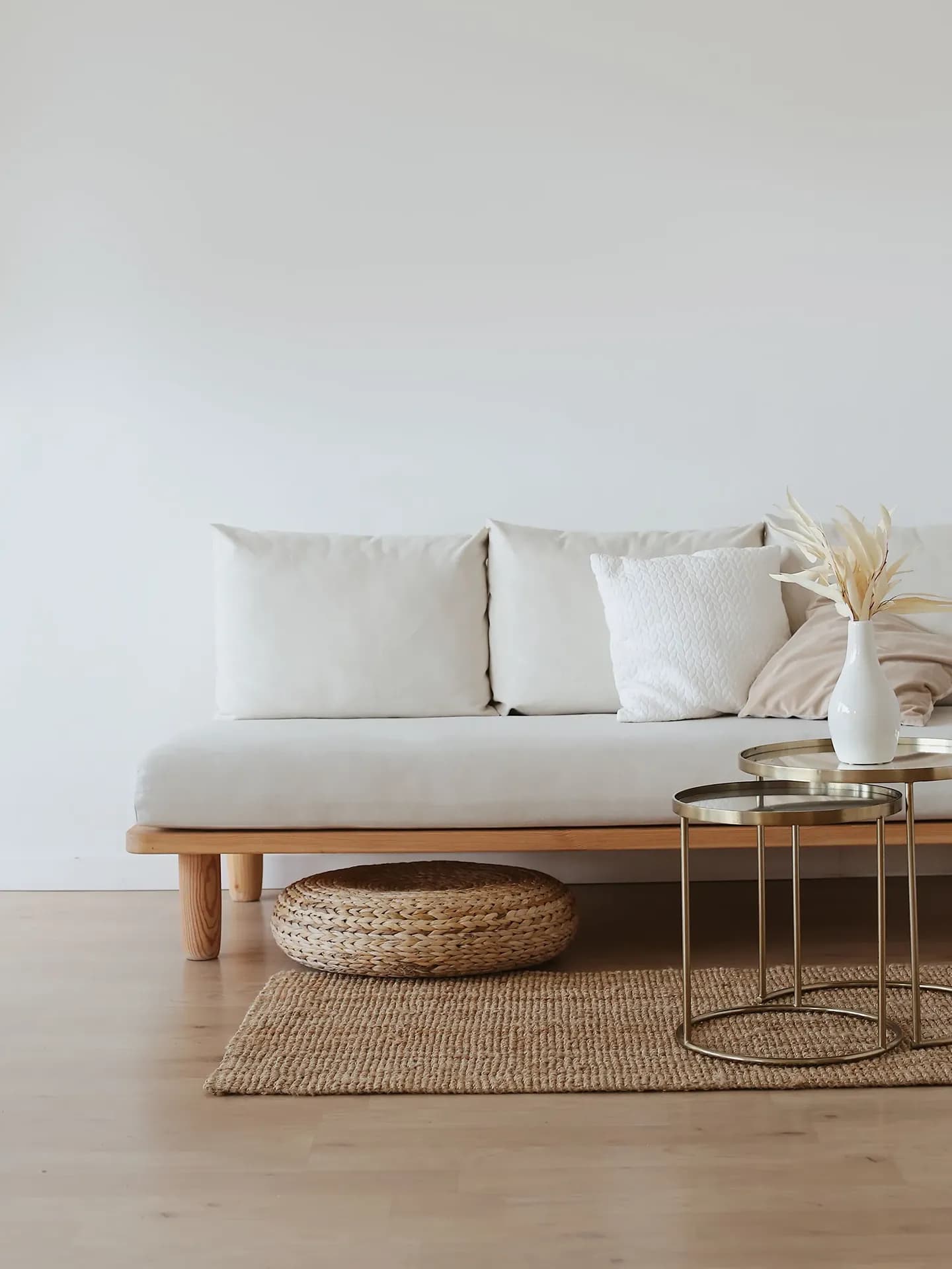 A bright, warm minimalist living room setup featuring a light-beige wooden sofa with cream-colored cushions, a woven jute rug, a round wicker pouf underneath, and tiered gold nesting coffee tables holding a white ceramic vase with dried pampas grass