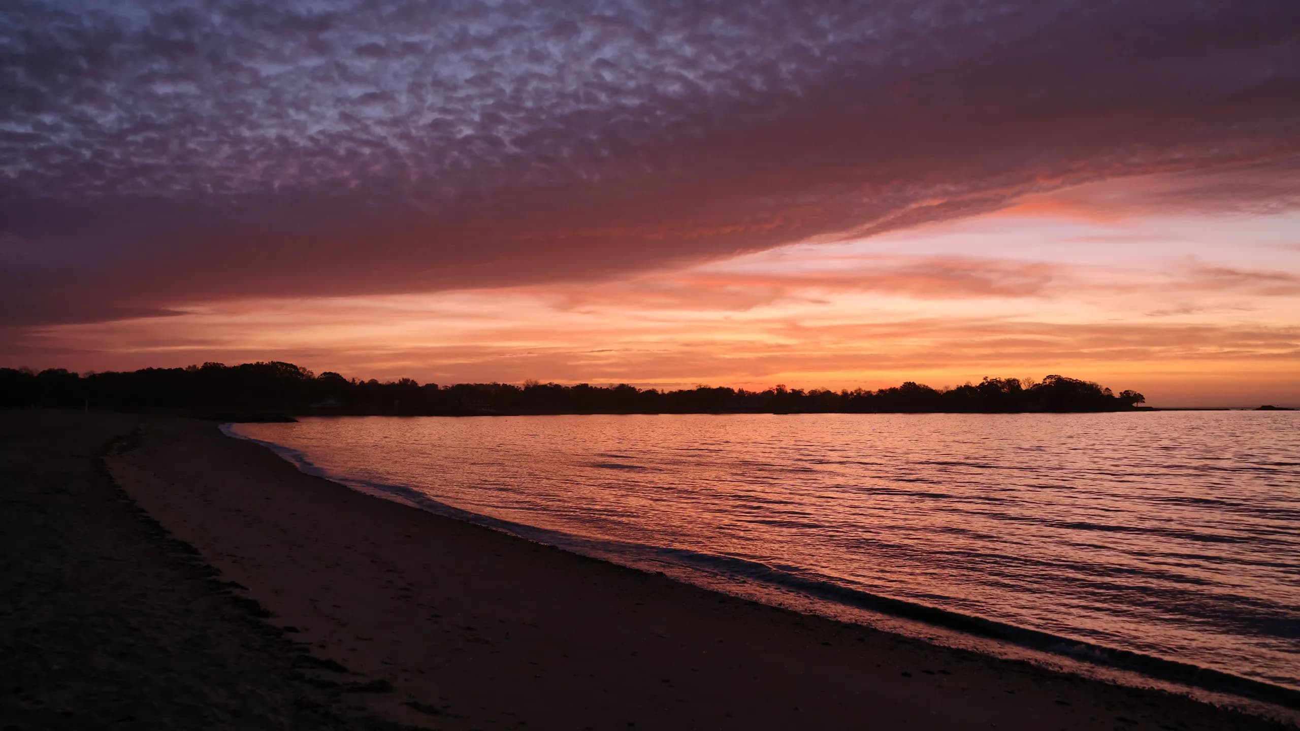 A serene view of a beach at sunset, with the sky covered in highly textured clouds painted in violet, pink, and bright orange reflecting over the rippling ocean surface, bordered by a dark shoreline.