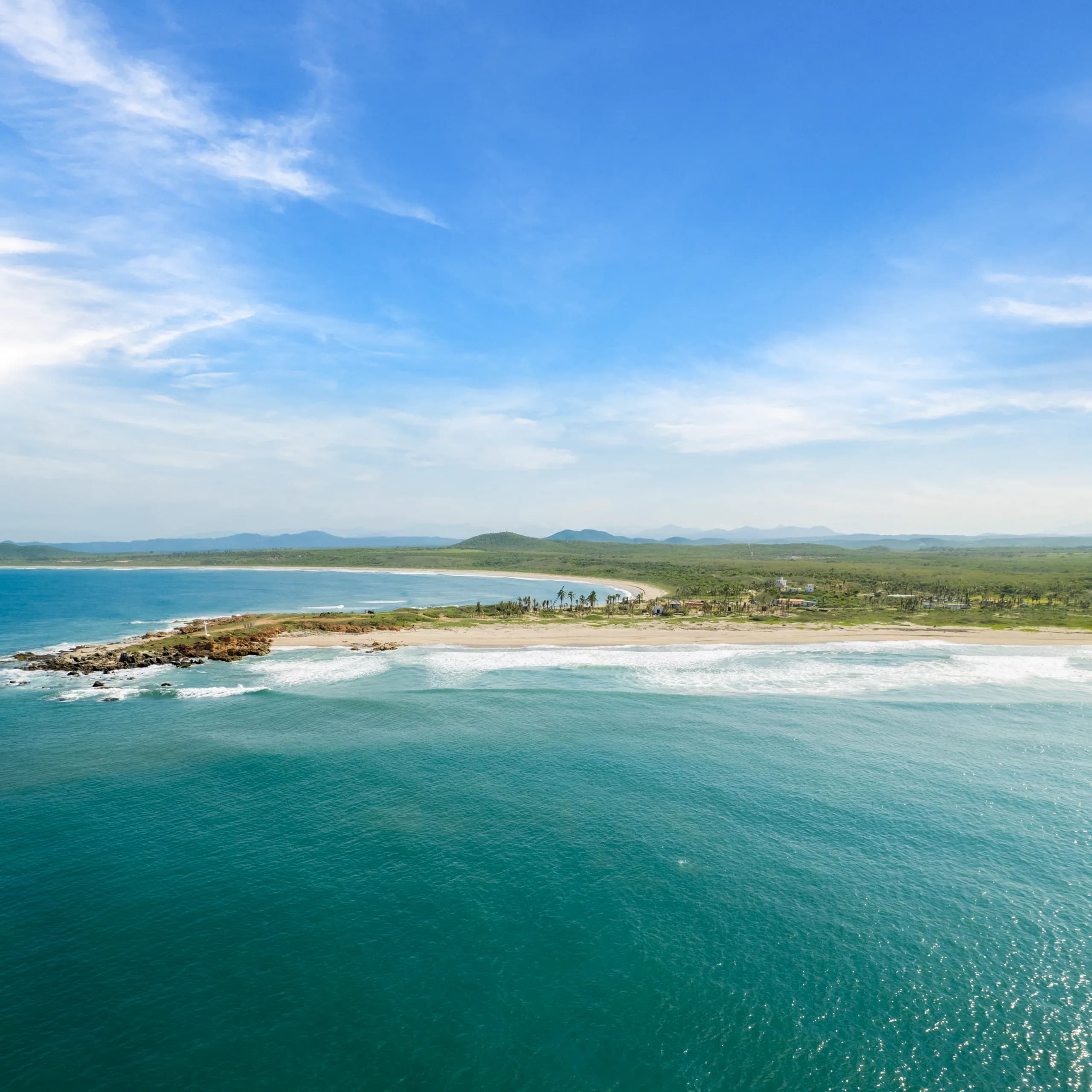 Panoramic aerial view of a rocky point and a pristine beach in Cabo Corrientes. The ocean features a deep turquoise hue with white waves, while the coastline extends toward a green plain and low hills under a bright blue sky with light clouds.