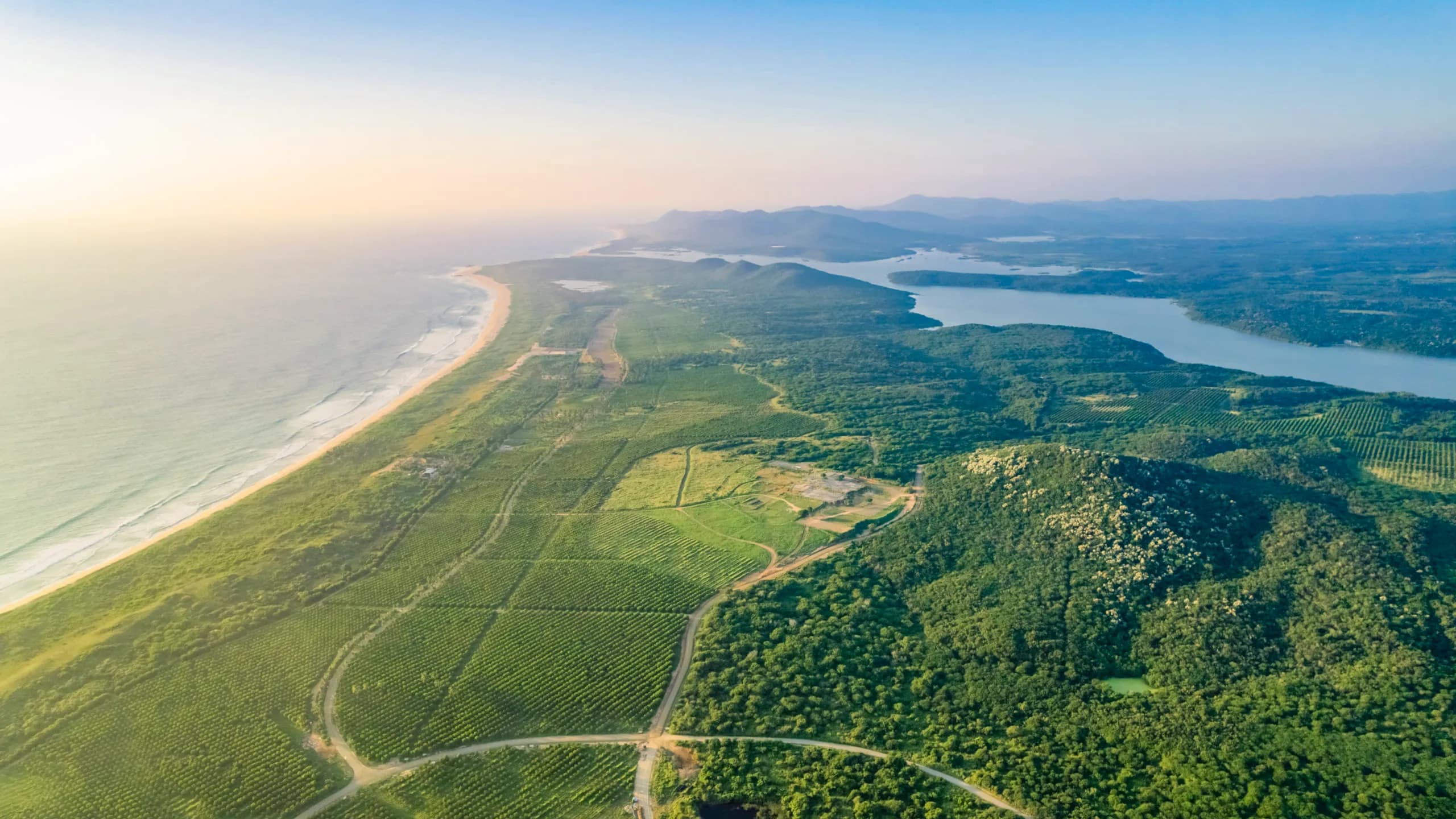 Panoramic aerial view of the Xala area, showing an extensive peninsula of green land and agricultural fields bordered on the left by a long golden sand beach with rolling waves, and on the right by a large lagoon or estuary. Forested hills extend toward the horizon under a soft sunset light.