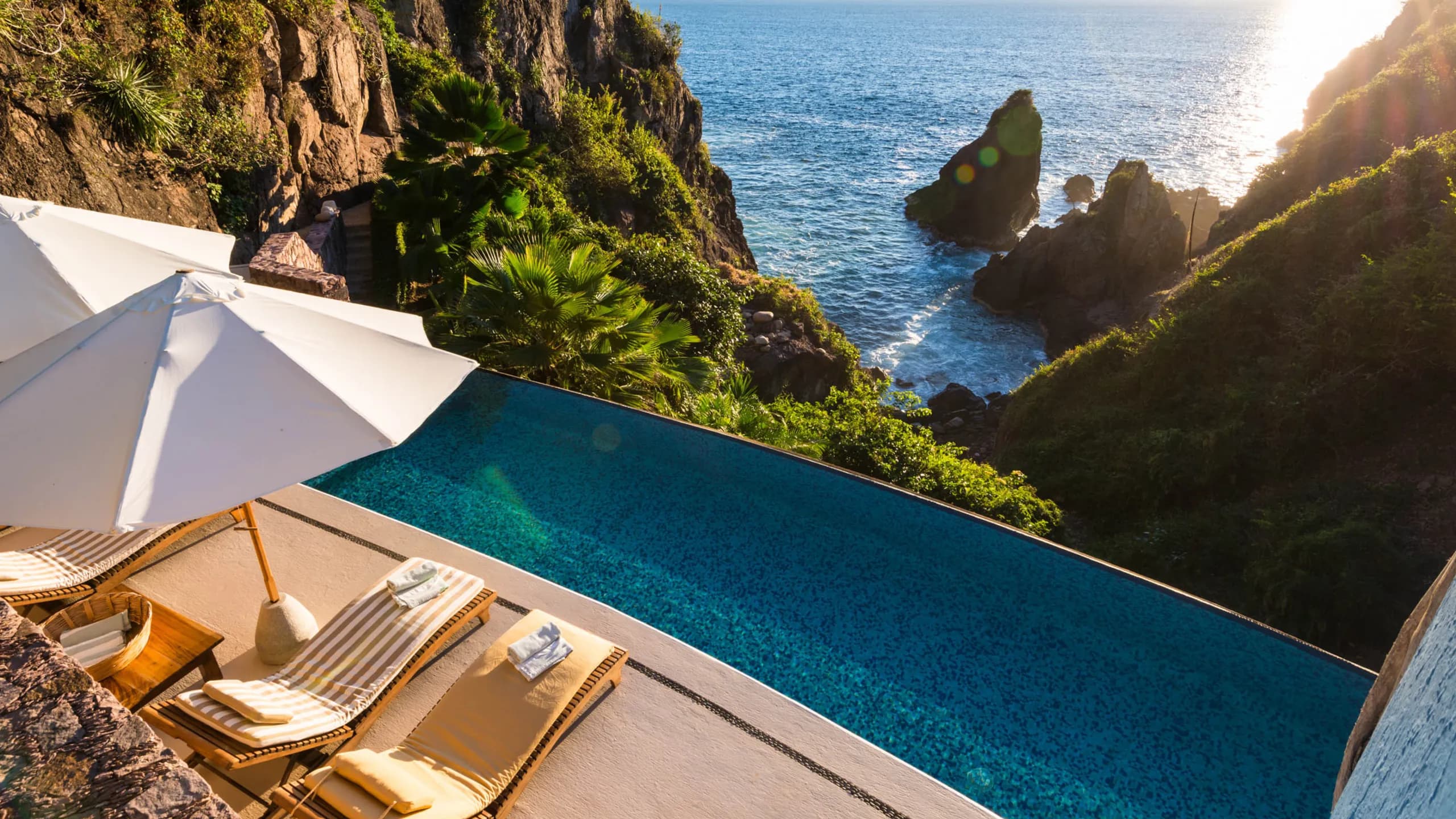 Elevated view of a rectangular infinity pool embedded into a steep cliffside. On the terrace are beige-striped lounge chairs and white umbrellas. In the background, the pool water visually merges with the open sea breaking against large rocky formations covered in wild vegetation under the sunlight.