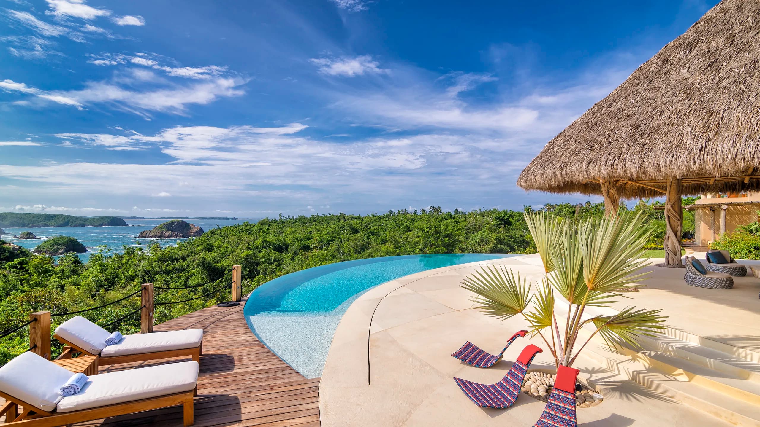A luxury terrace featuring a curved infinity pool that blends into the landscape. On the right, a palapa with rustic wooden columns and light-colored stone flooring; on the left, a wooden deck with white lounge chairs. In the background, a dense green jungle, rocky islets in the blue sea, and a bright sky with scattered clouds.