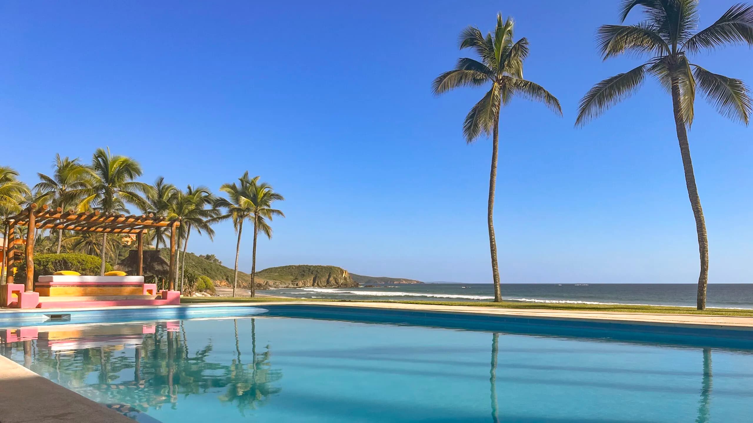 A large pool of crystal-clear blue water facing the ocean in Careyes. In the foreground to the left, a lounge area with a wooden pergola and pink and yellow accents; to the right, two tall, slender palm trees rise against a clear blue sky, with gentle waves breaking on the beach in the background.