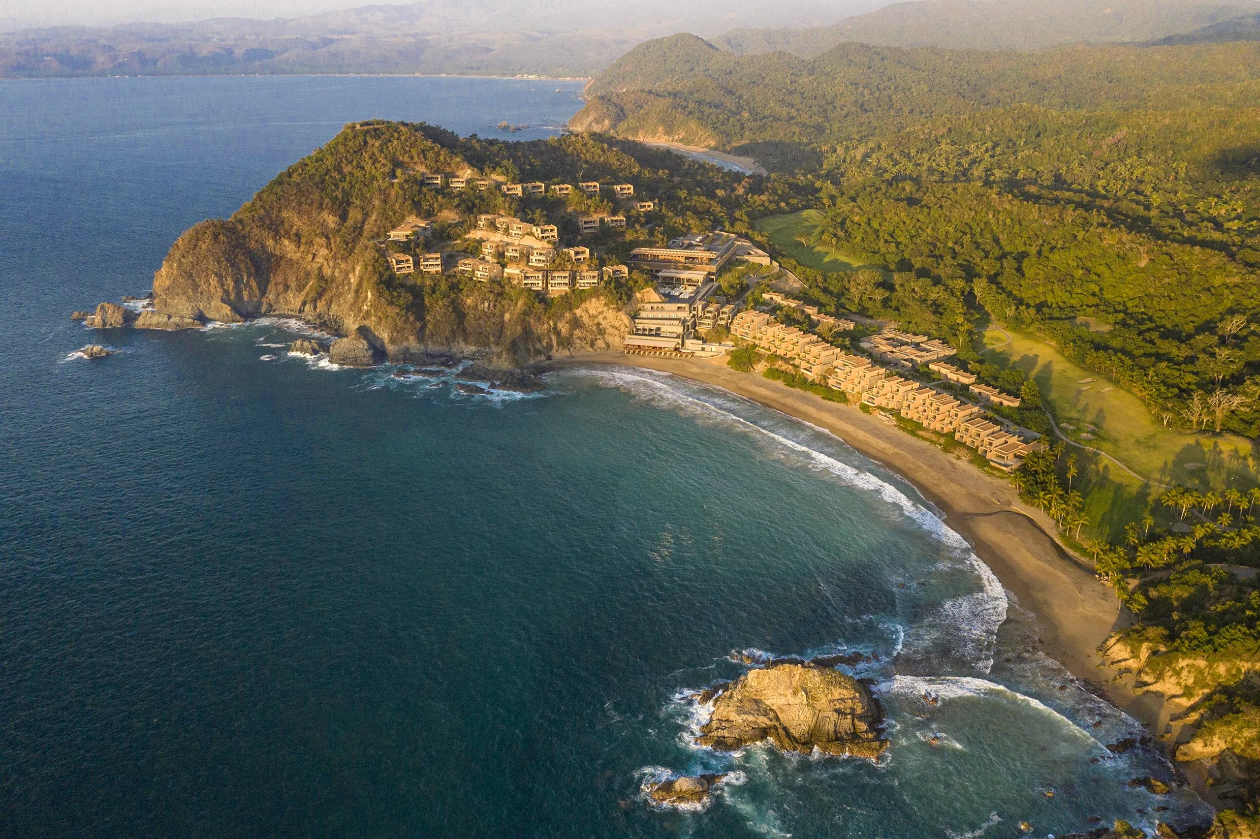 Aerial view of the Four Seasons Tamarindo hotel integrated into a cliff covered in dense tropical vegetation. The architectural complex descends in earth-toned terraces toward a crescent-shaped private beach, washed by a deep blue sea with rocky islets off the coast.