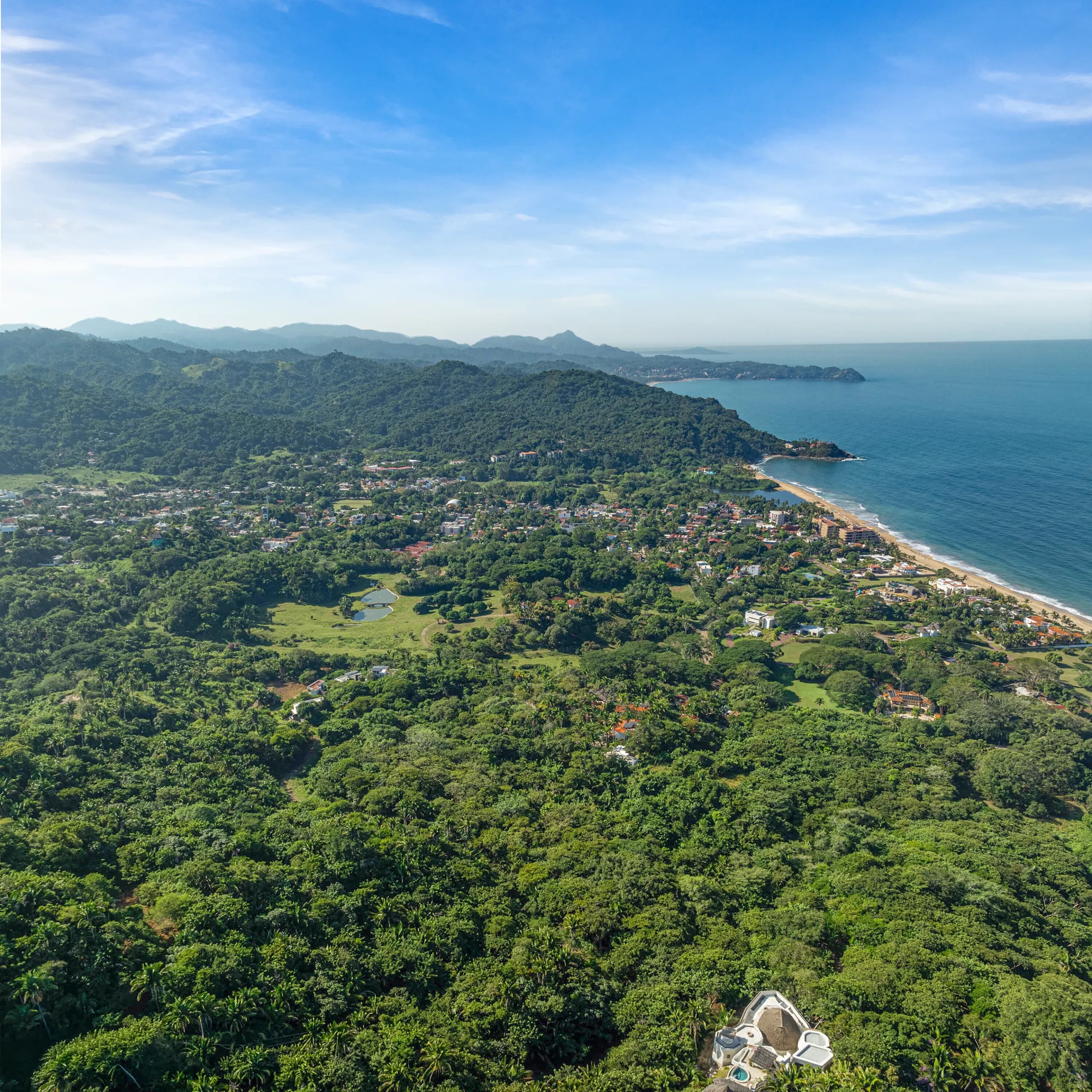 Panoramic aerial view of San Pancho, showing a vast, lush green jungle canopy rolling down to meet a long sandy beach coastline and the blue Pacific ocean under a bright sky.