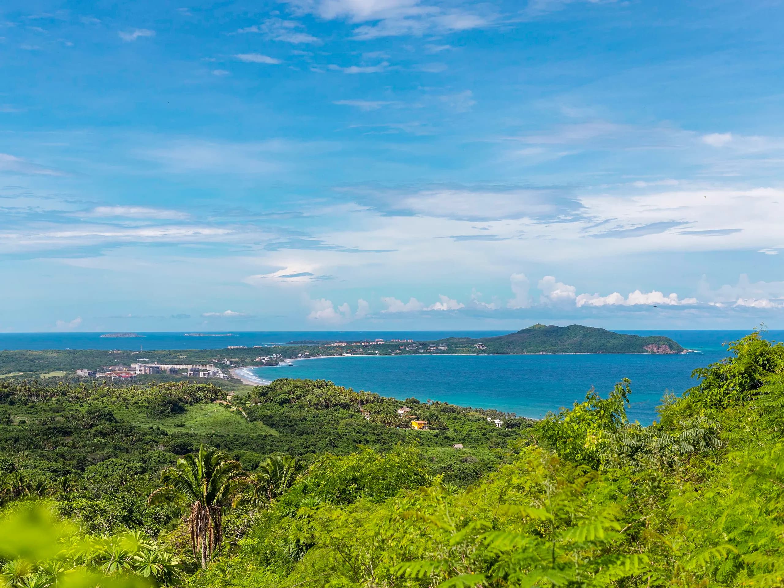Panoramic elevated view of a crescent-shaped bay along the Riviera Nayarit coastline, featuring lush green forested hills and distant resorts meeting the blue ocean under a bright sky in Higuera Blanca.