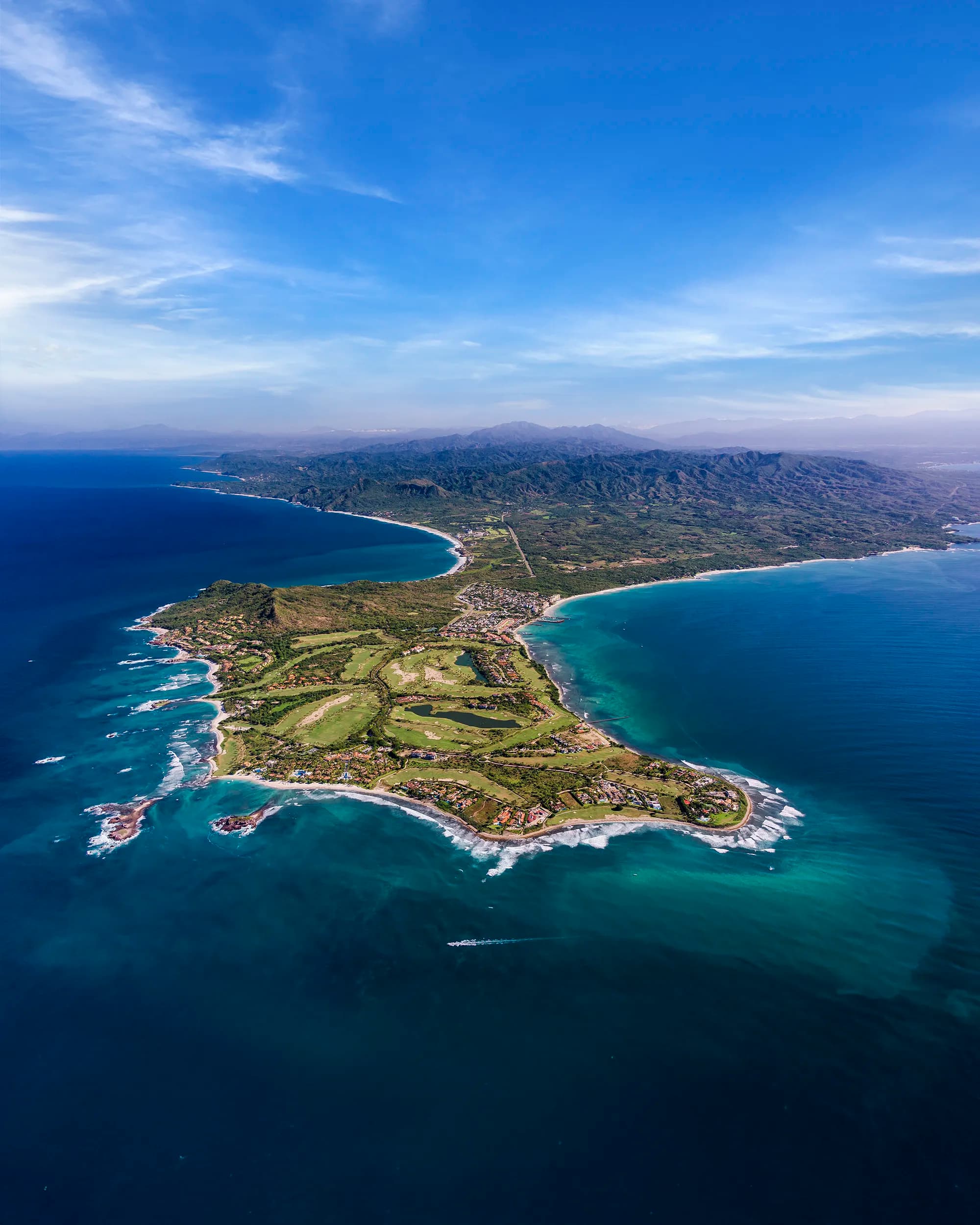 Breathtaking high-altitude aerial view of the Punta Mita peninsula, showing its signature golf courses and luxury properties surrounded by turquoise ocean waters, with the northern Riviera Nayarit coastal corridor extending into the distant mountains.
