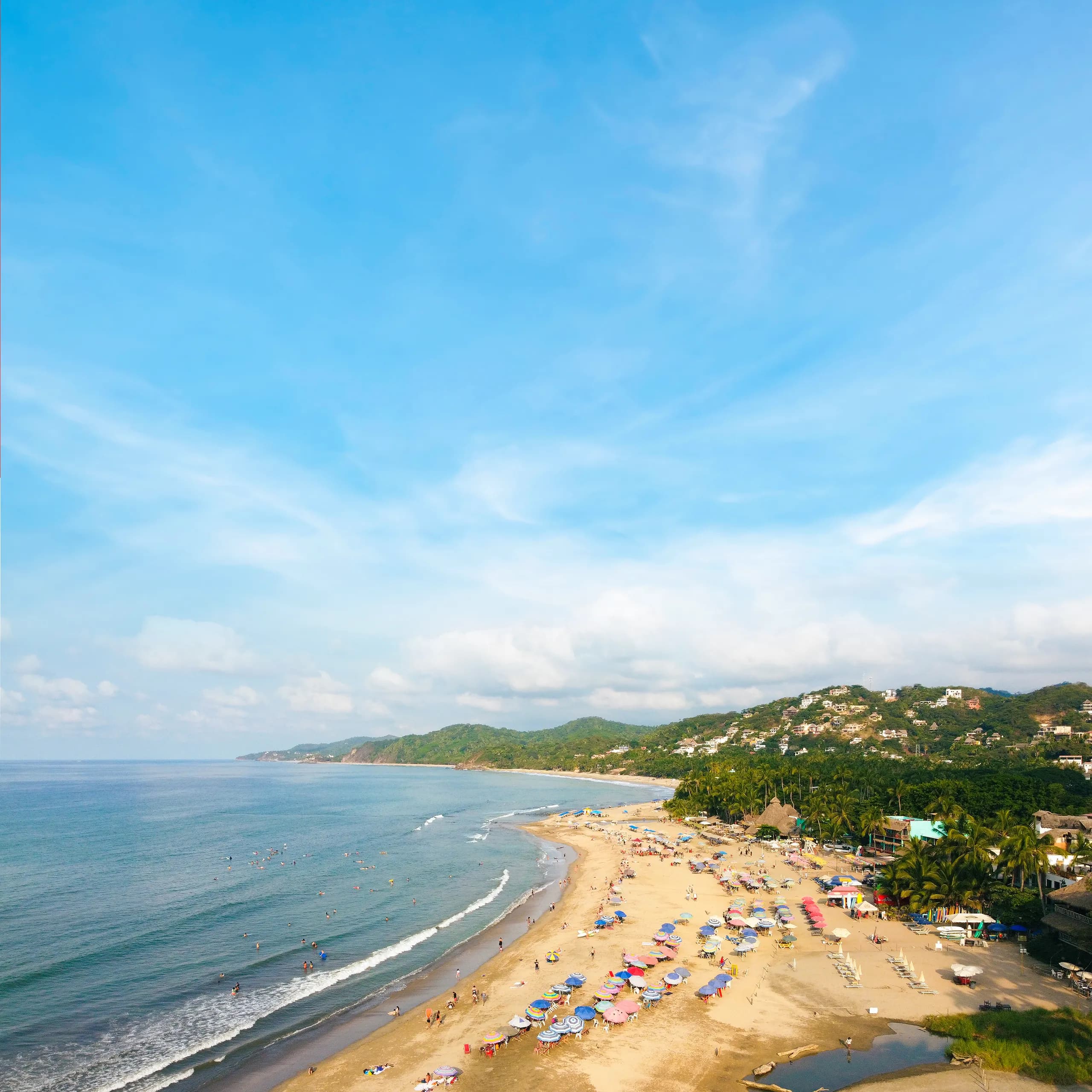 High-angle view of Sayulita beach, showing a wide sandy shore lined with colorful umbrellas and beachgoers, bordering a blue ocean bay with surfers and green coastal hills under a partly cloudy sky.