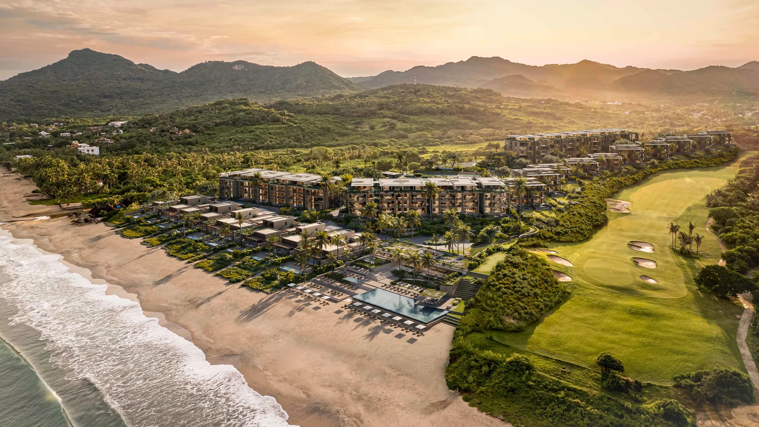 Elevated view of Litibú Bay Club featuring luxury beachfront residences, an infinity pool, and an adjacent golf course nestled between green hills and the Pacific ocean at sunset.