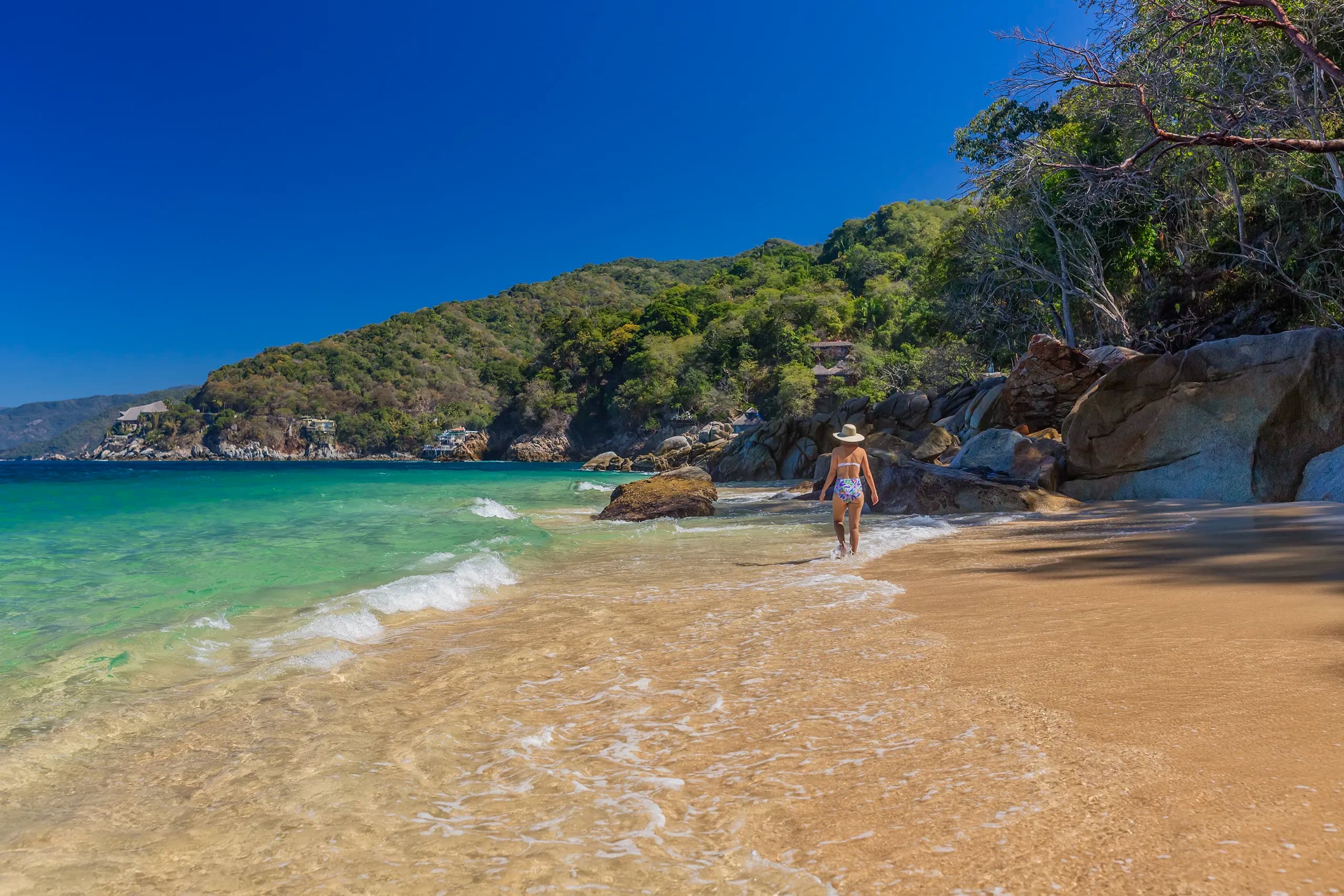 A woman wearing a sun hat walks along the shoreline of a tropical beach with clear turquoise water and golden sand. Lush, green jungle-covered hills rise up in the background under a bright blue sky.